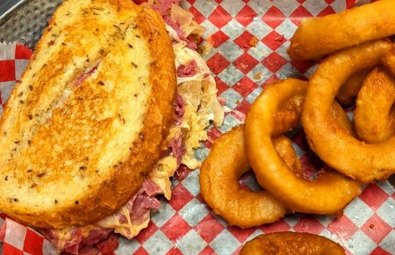 A sandwich with sliced toasted bread, filled with meat and sauerkraut, alongside a pile of crispy onion rings.