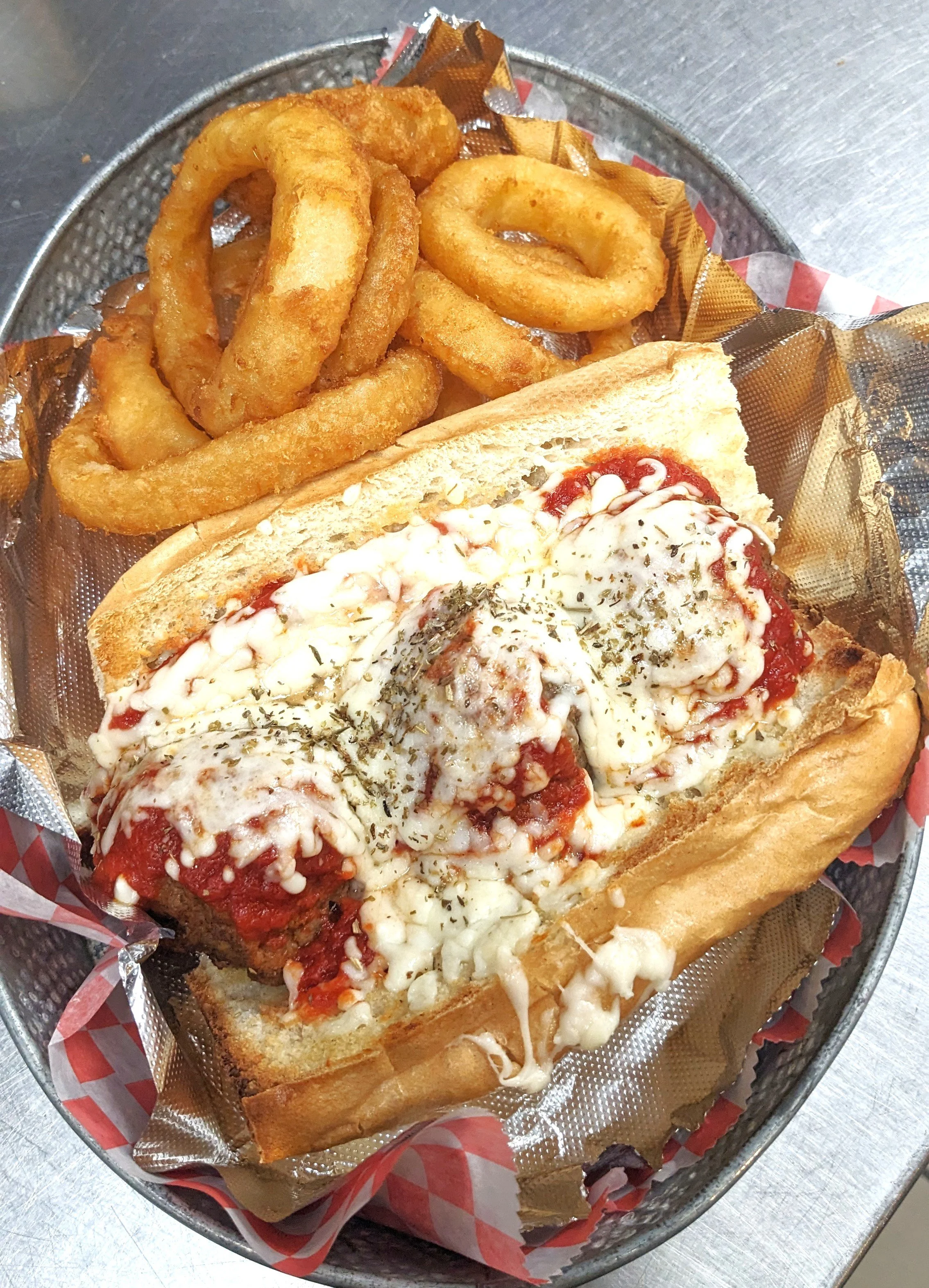 A basket containing a Philly cheesesteak sandwich topped with melted cheese and herbs, with curly fried onion rings on the side.