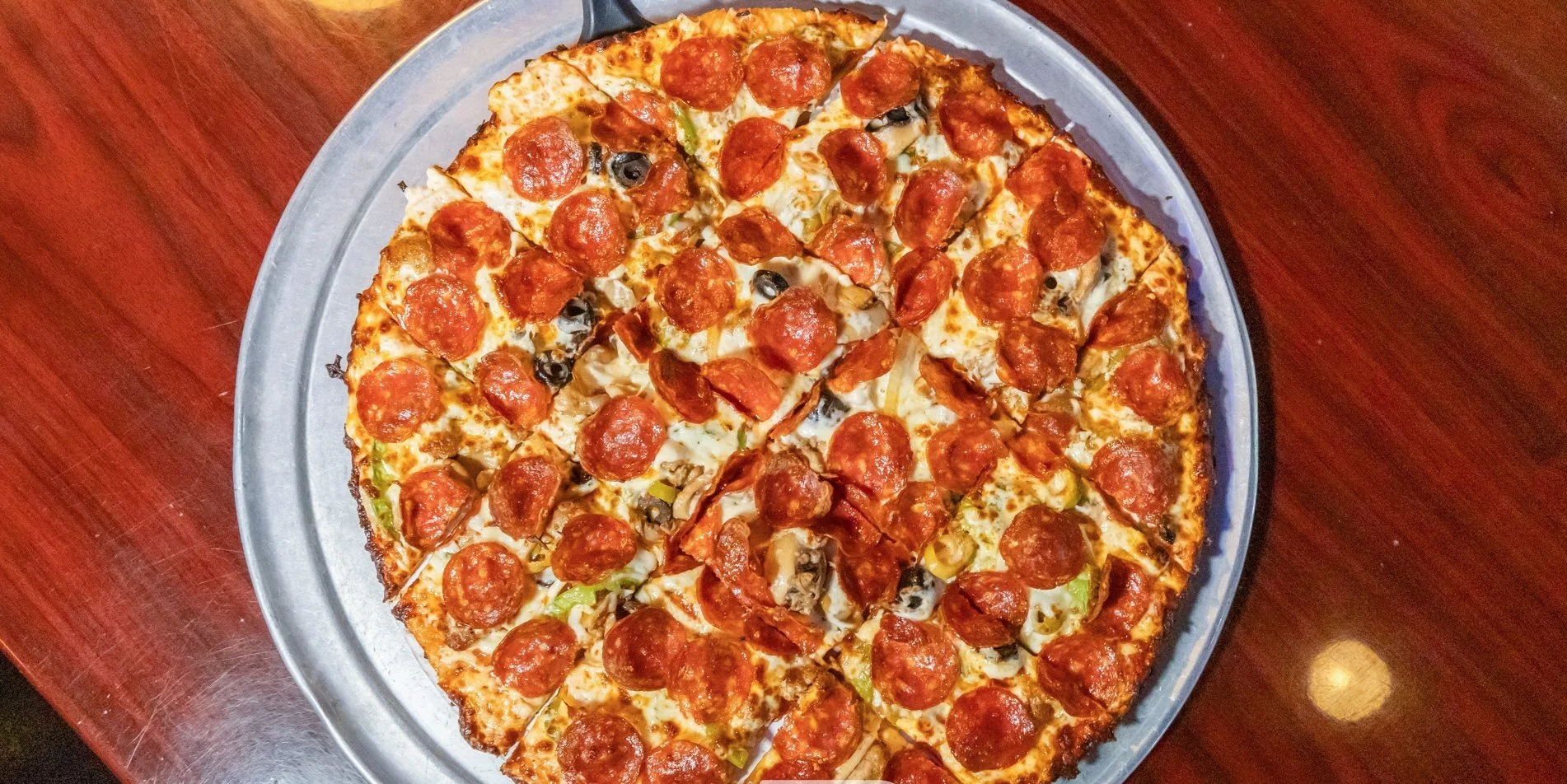 Pepperoni pizza with cheese, black olives, green peppers, and onions on a metal tray on a wooden table.