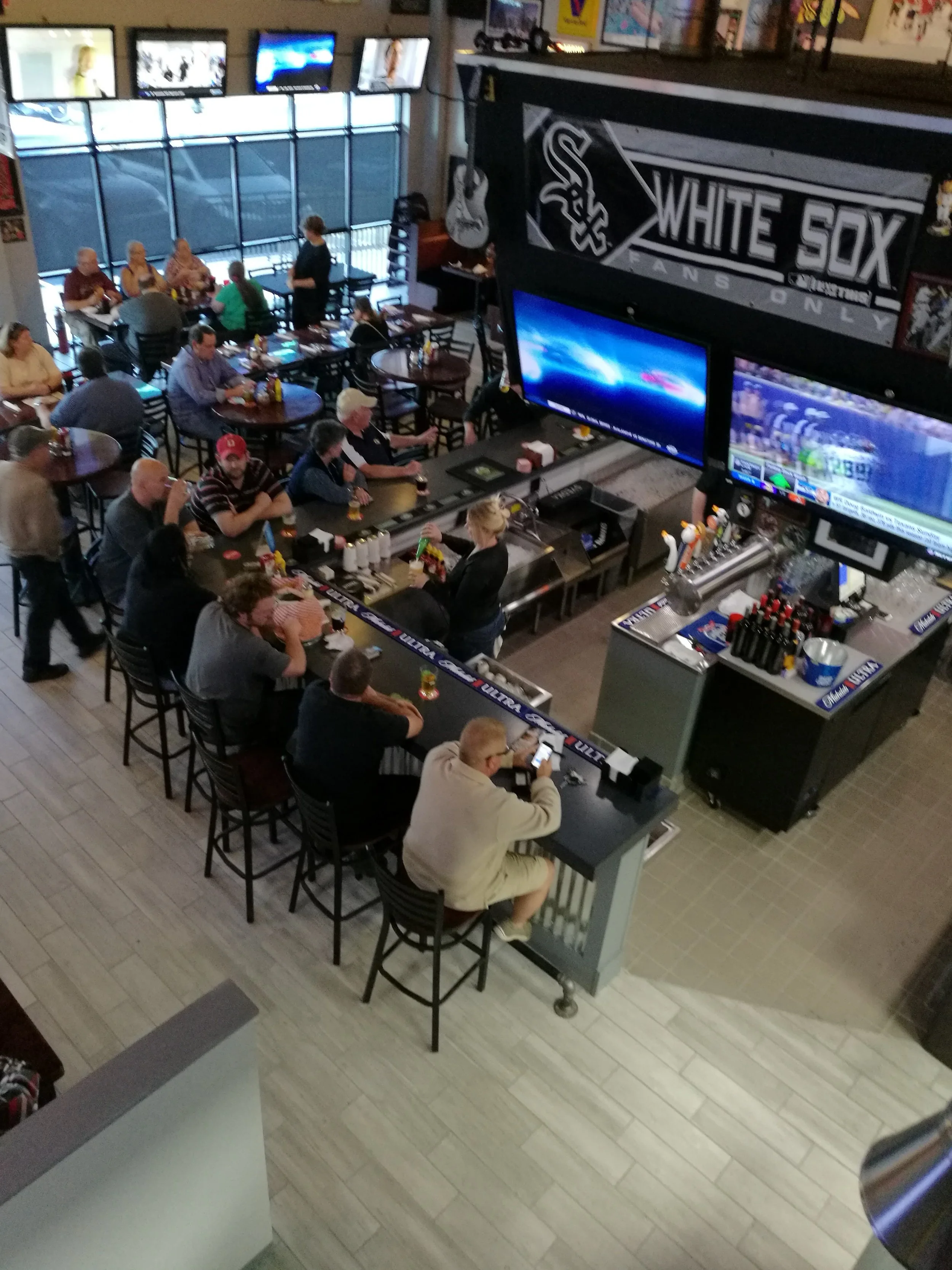 A sports bar filled with patrons, some sitting at tables and others at the bar, watching multiple large screens showing sports games and news.