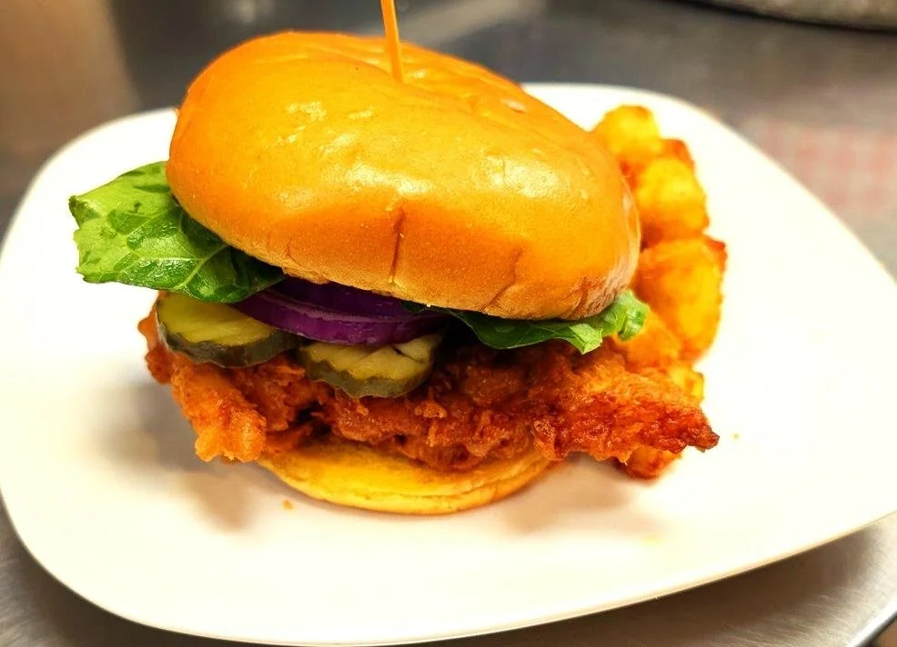 Fried chicken sandwich with pickles, lettuce, and red onion on a bun, served with curly fries on a white plate.