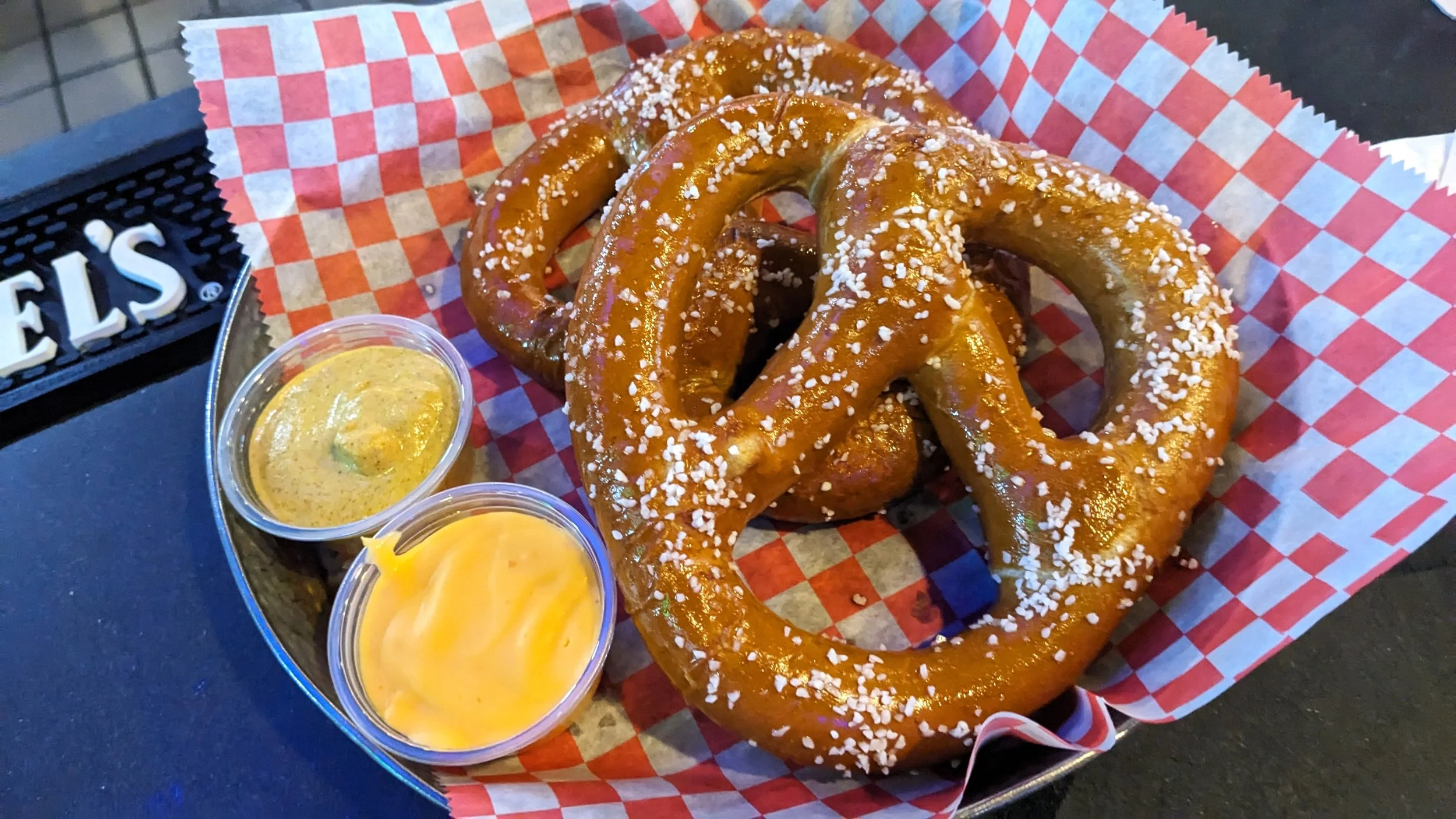 Three large salted pretzels on red and white checkered paper, served with two containers of melted cheese and mustard sauce.