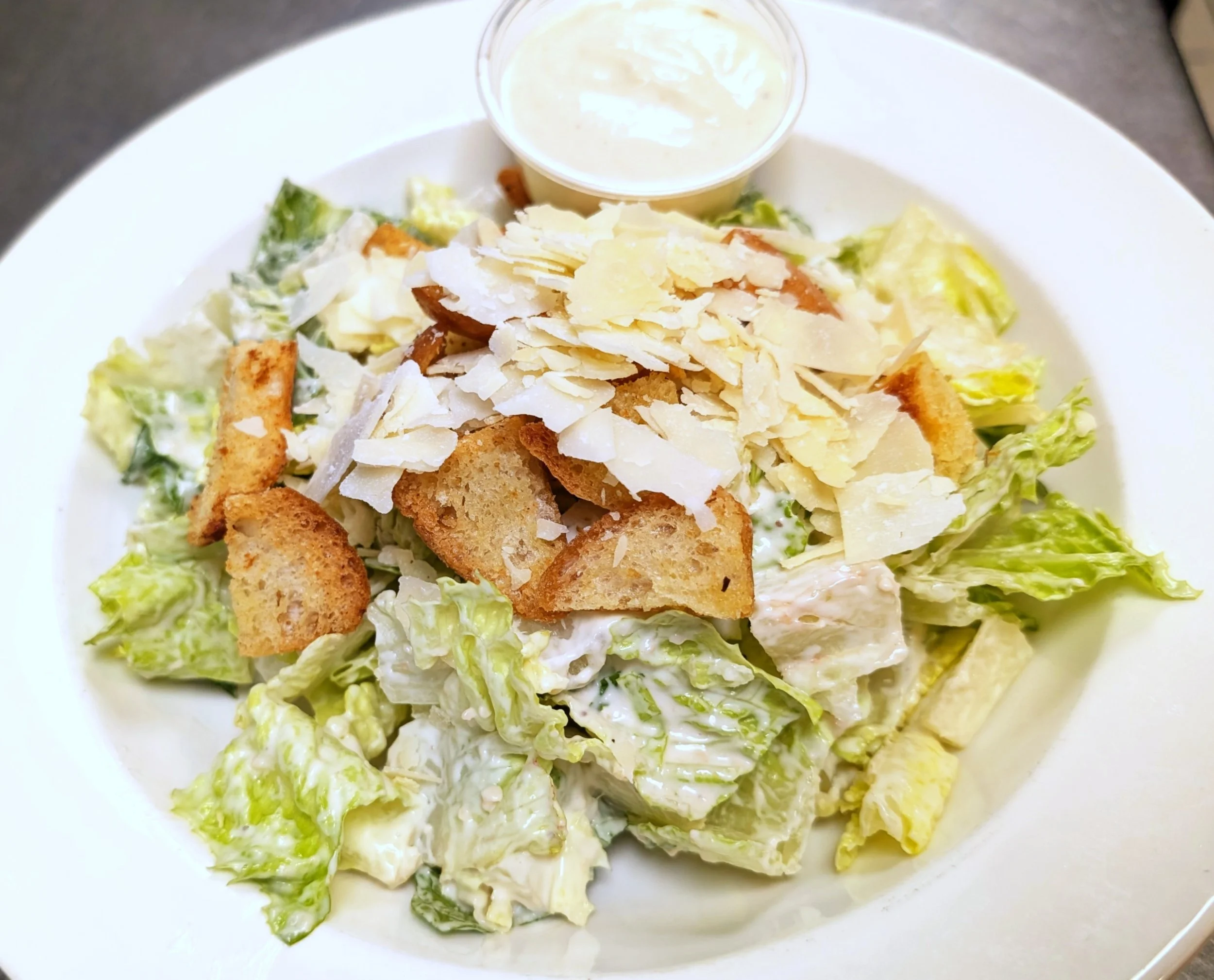 A Caesar salad topped with shredded Parmesan cheese and croutons, accompanied by a small cup of creamy dressing on a white plate.