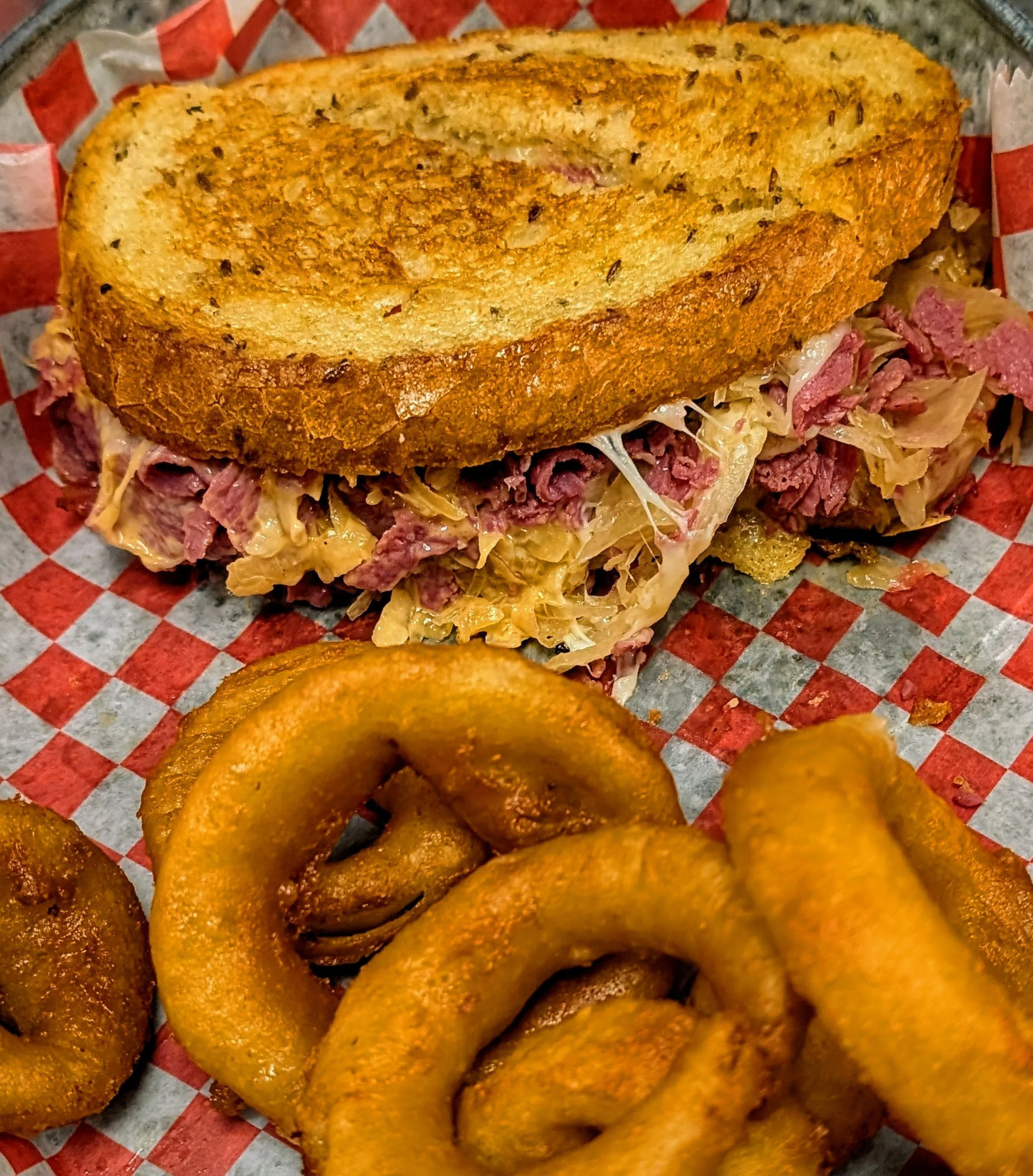 A Reuben sandwich with rye bread, corned beef, sauerkraut, and melted cheese, served with a side of onion rings.