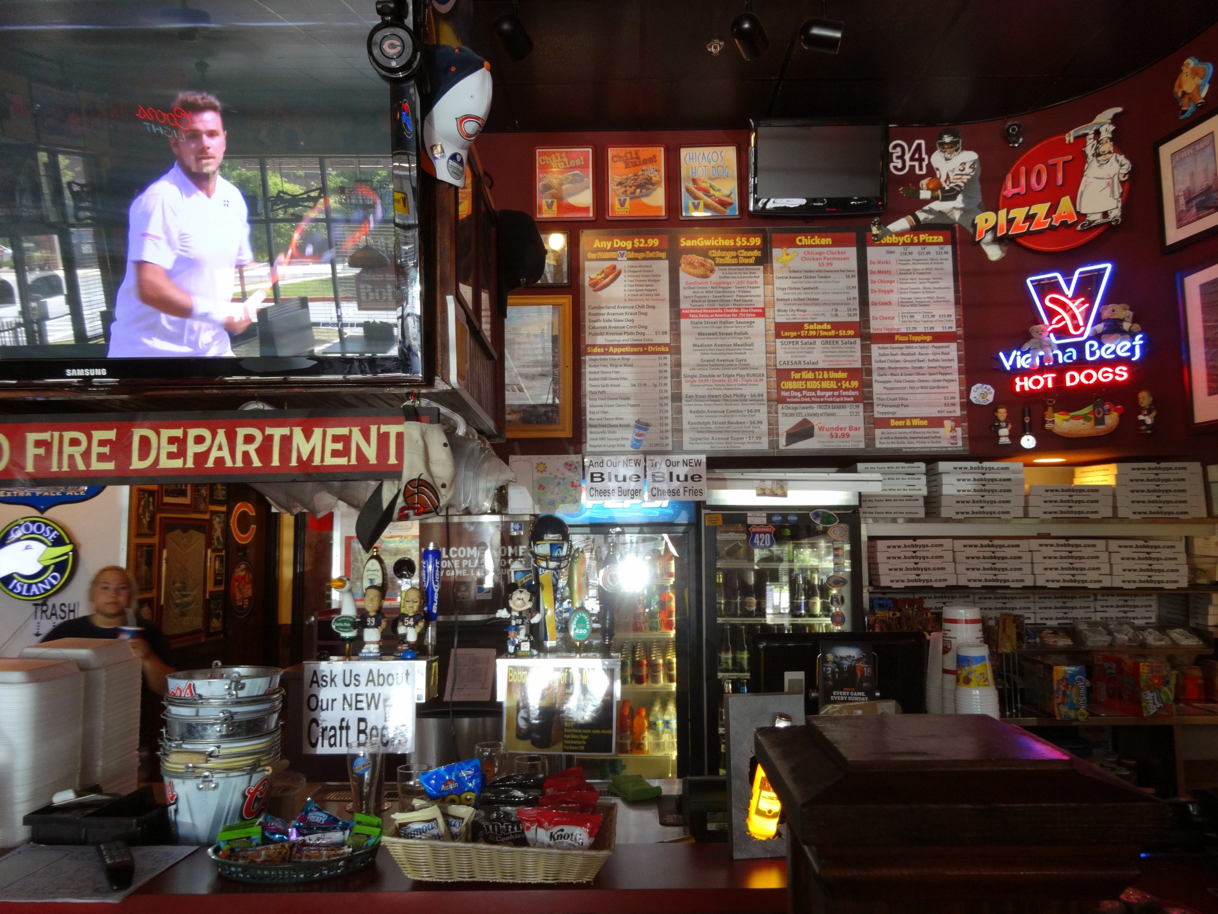 Interior of a pizza restaurant with menu boards, neon signs, a television showing a man in a chef's coat, a captain's hat, and various decorations and snacks. A staff member is visible behind the counter.