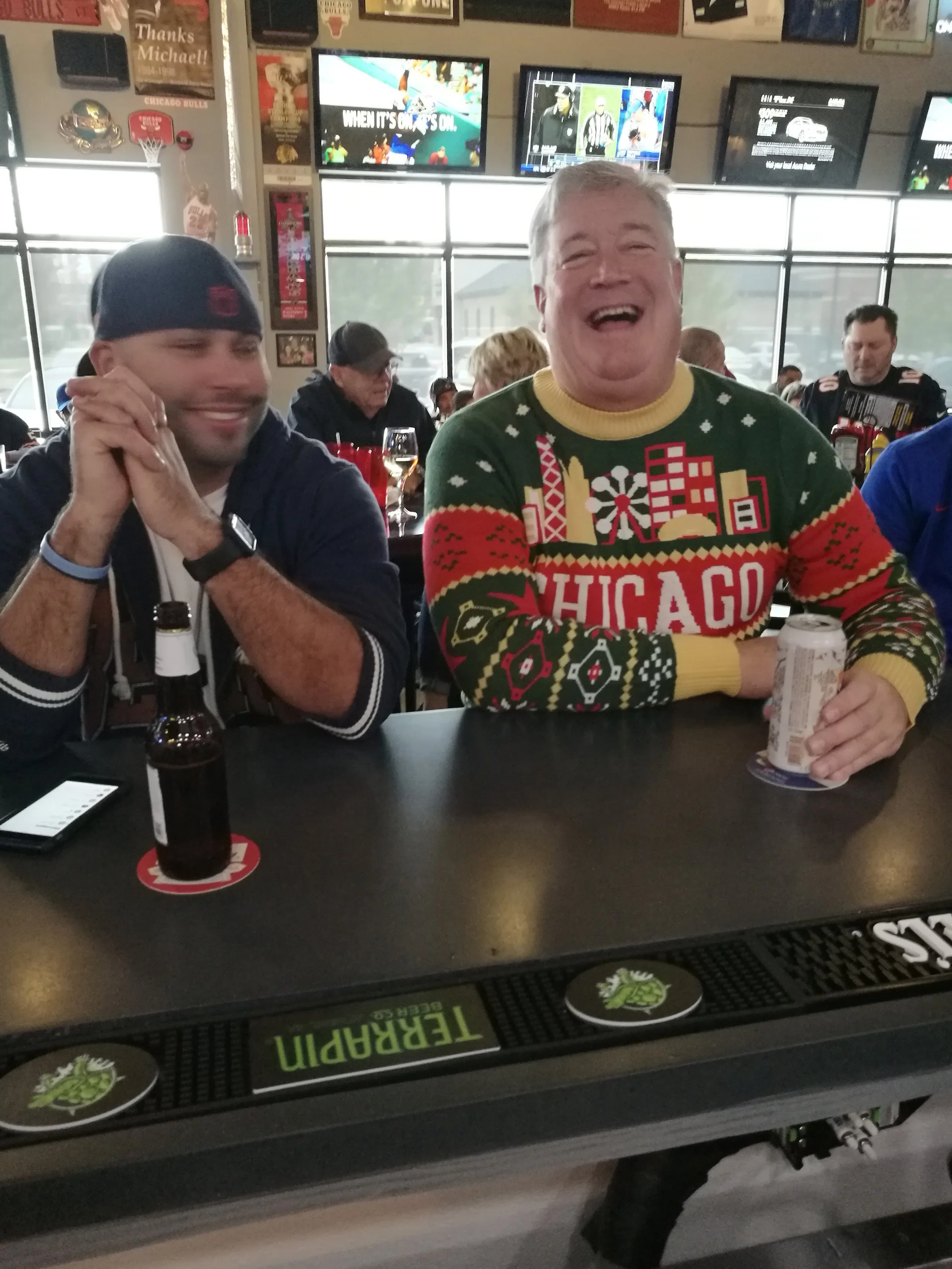 Two men sitting at a bar, one smiling broadly wearing a green sweater with a Chicago theme, and the other with hands clasped smiling, with drinks and a smartphone on the bar in front of them, in a lively sports bar setting.