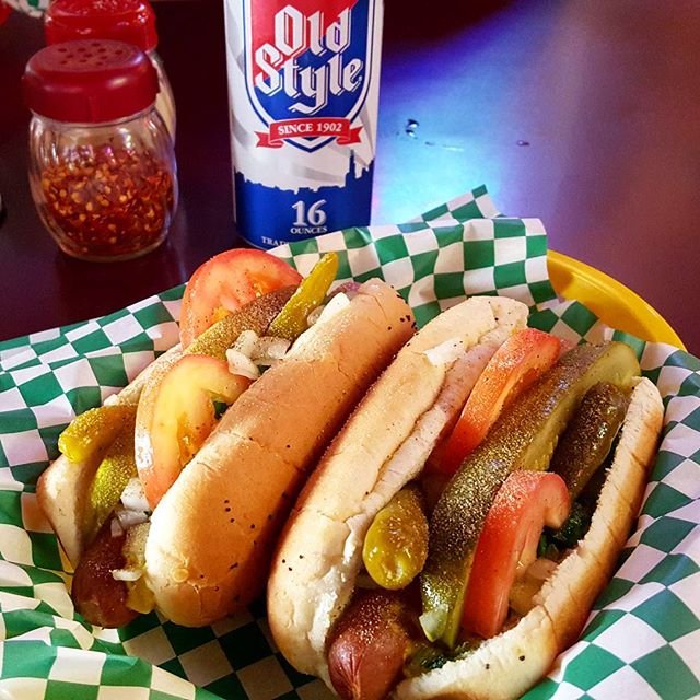 Two hot dogs topped with pickles, tomatoes, and onions in a basket with green and white checkered paper, and a can of Old Style beer and a jar of crushed red pepper on a dark table.