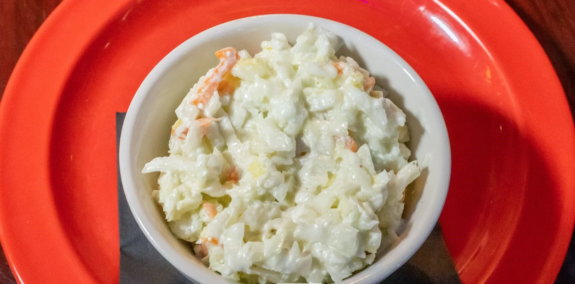 A dish of creamy potato salad with chopped vegetables in a white bowl on an orange plate.