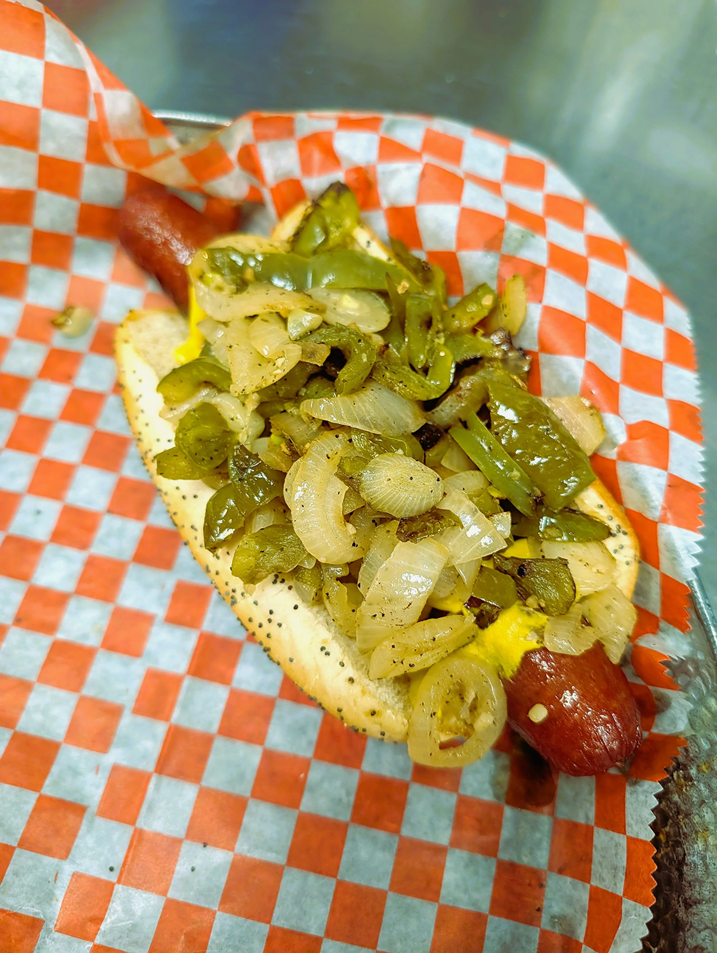 Hot dog with chopped grilled onions and green peppers on a bun, served on checkered paper in a metal tray.