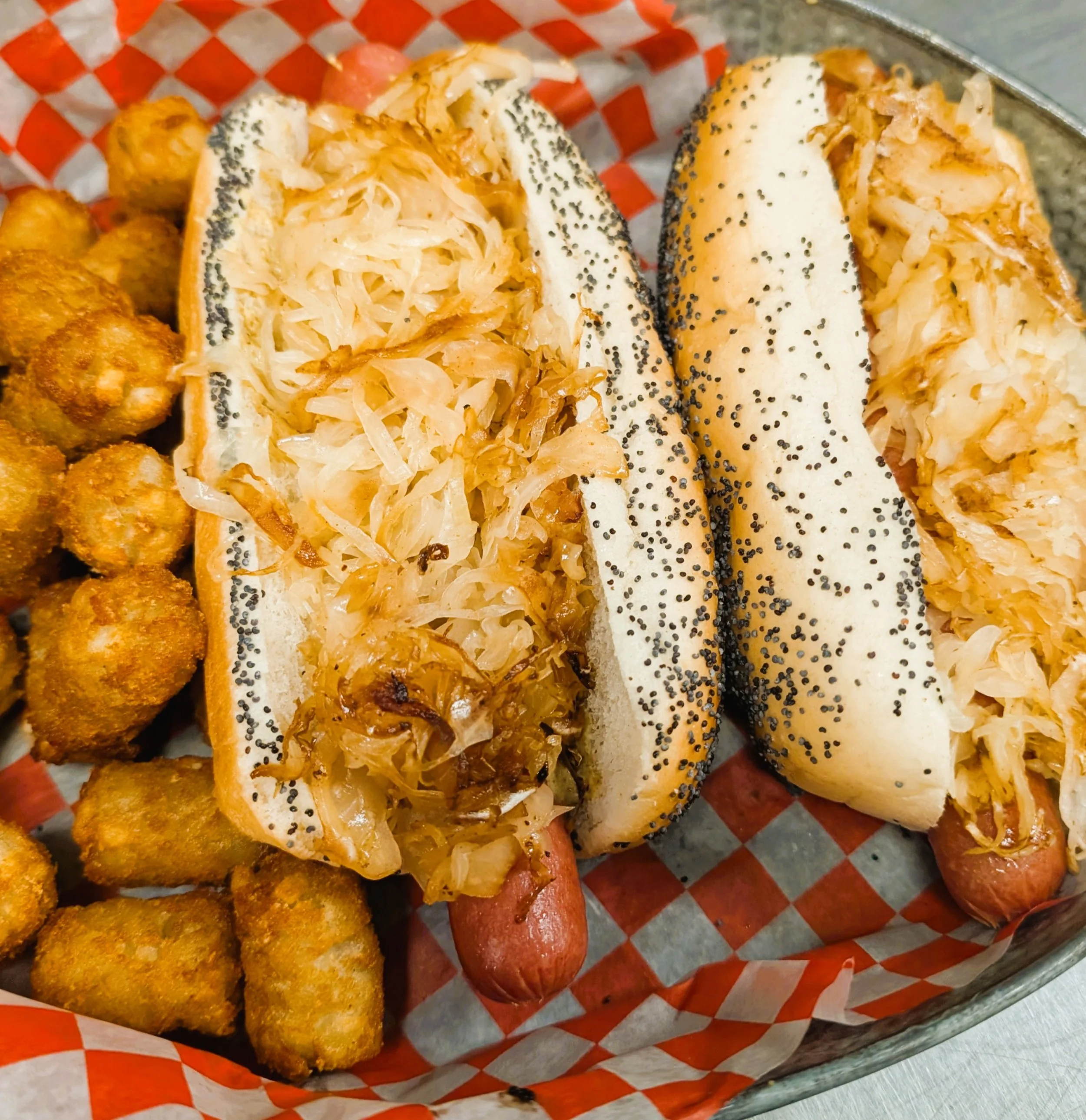 Close-up of two hot dogs with sauerkraut in poppy seed buns, served with fried tater tots on a red and white checkered paper-lined tray.