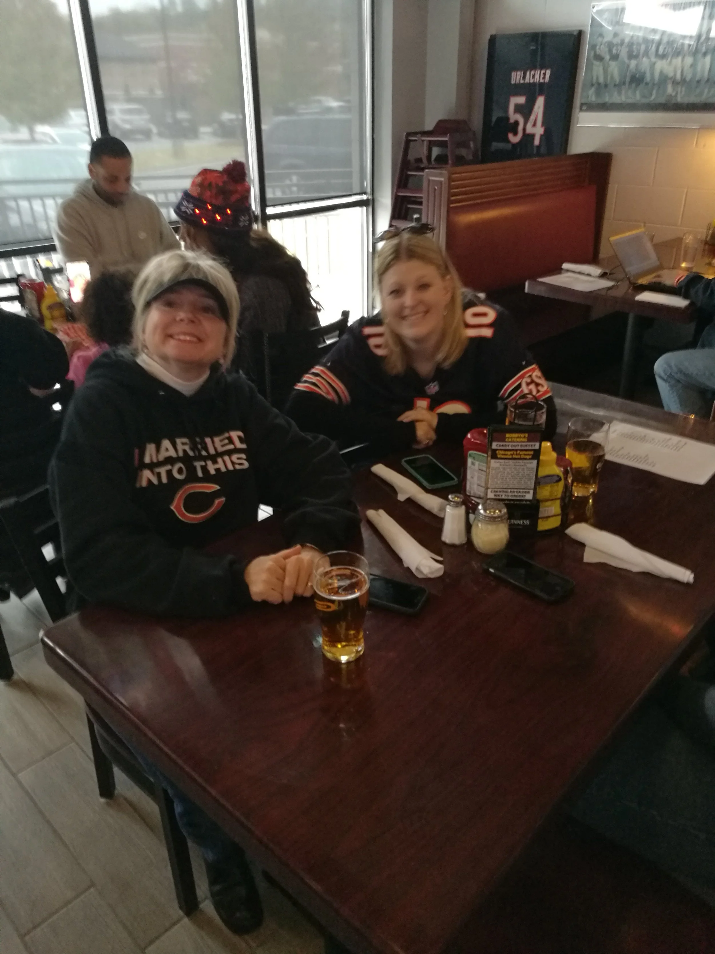 Two women in Chicago Bears jerseys sitting at a restaurant table with drinks and condiments, smiling at the camera.
