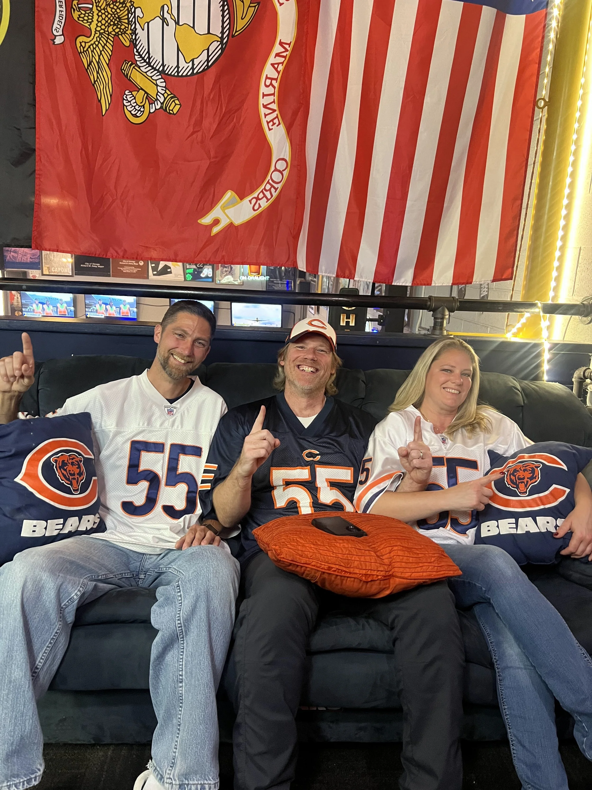 Three football fans sitting on a couch with Chicago Bears themed pillows, wearing Chicago Bears jerseys, and showing their team spirit. A large Chicago Bears flag hangs behind them.