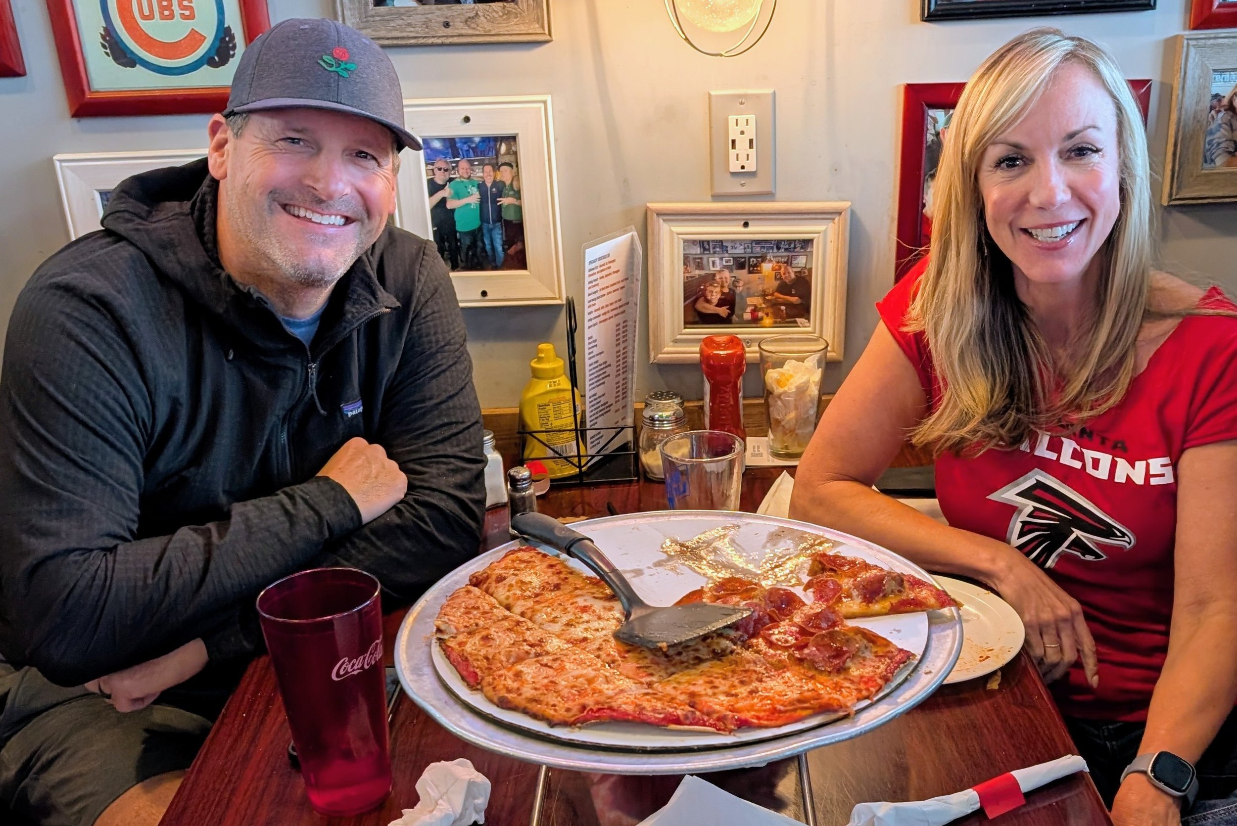 Man and woman smiling at a restaurant table with pizza, drink glasses, and condiments.
