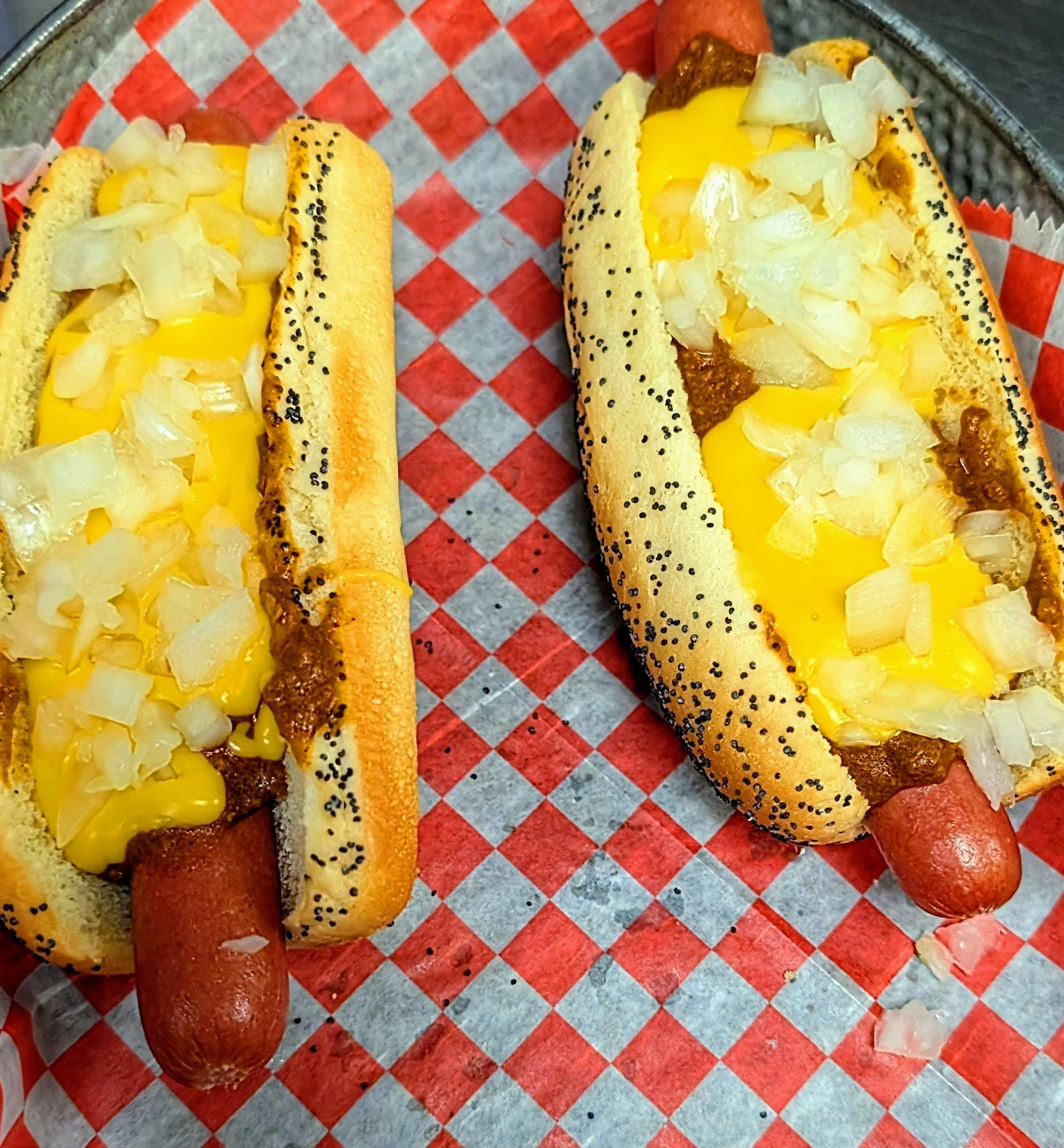 Two hot dog sandwiches with poppy seed buns, sausages, topped with yellow mustard and chopped onions, on a red checkered paper-lined tray.