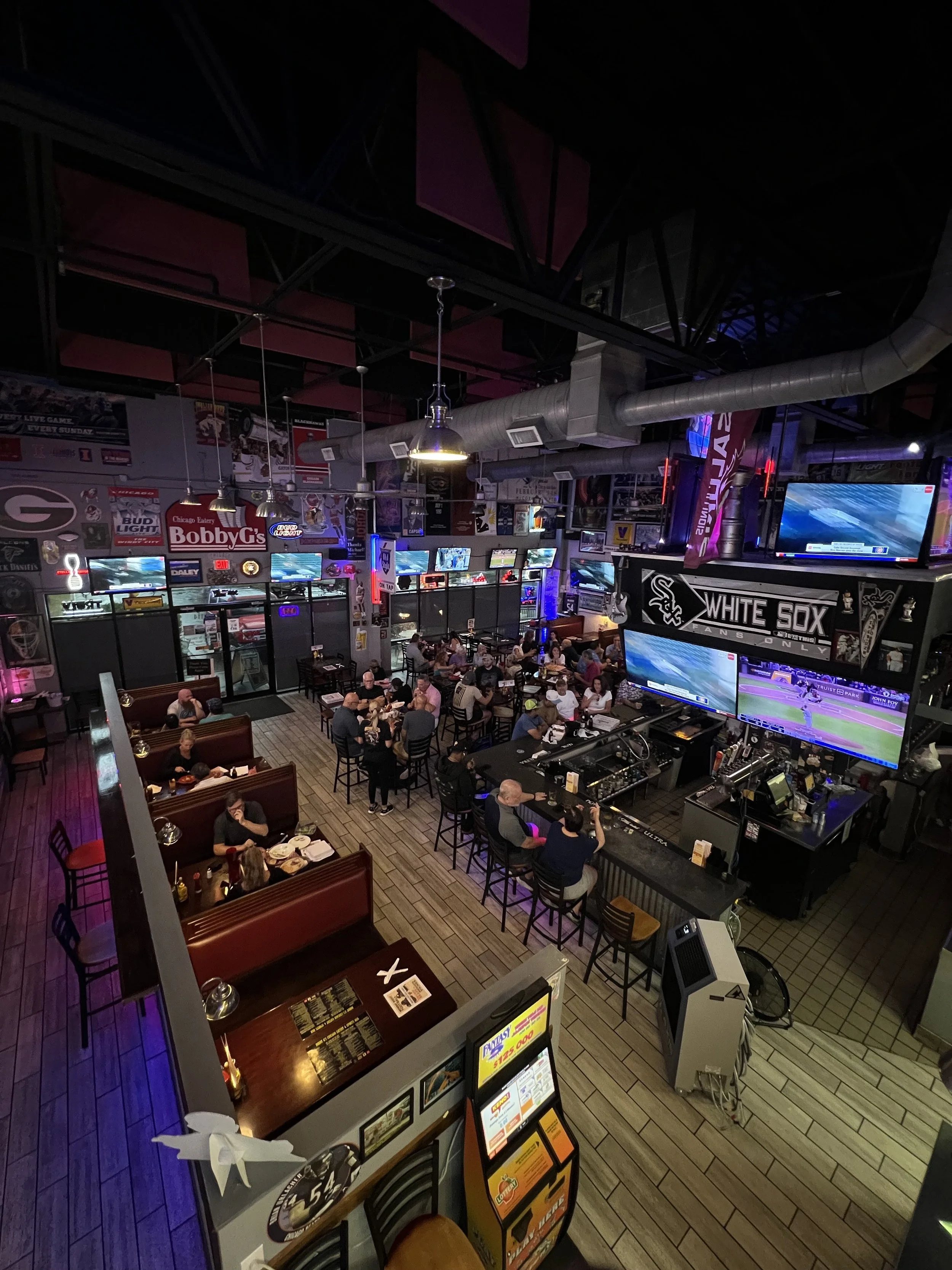 A lively sports bar with multiple TVs showing a baseball game, patrons seated at booths and tables enjoying food and drinks, illuminated with vibrant lighting and sporting memorabilia on the walls.