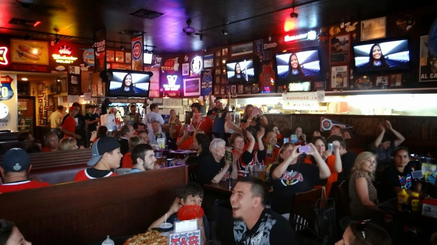 Group of people watching a sports game at a bar or restaurant, with multiple TV screens, neon signs, and vintage decor.