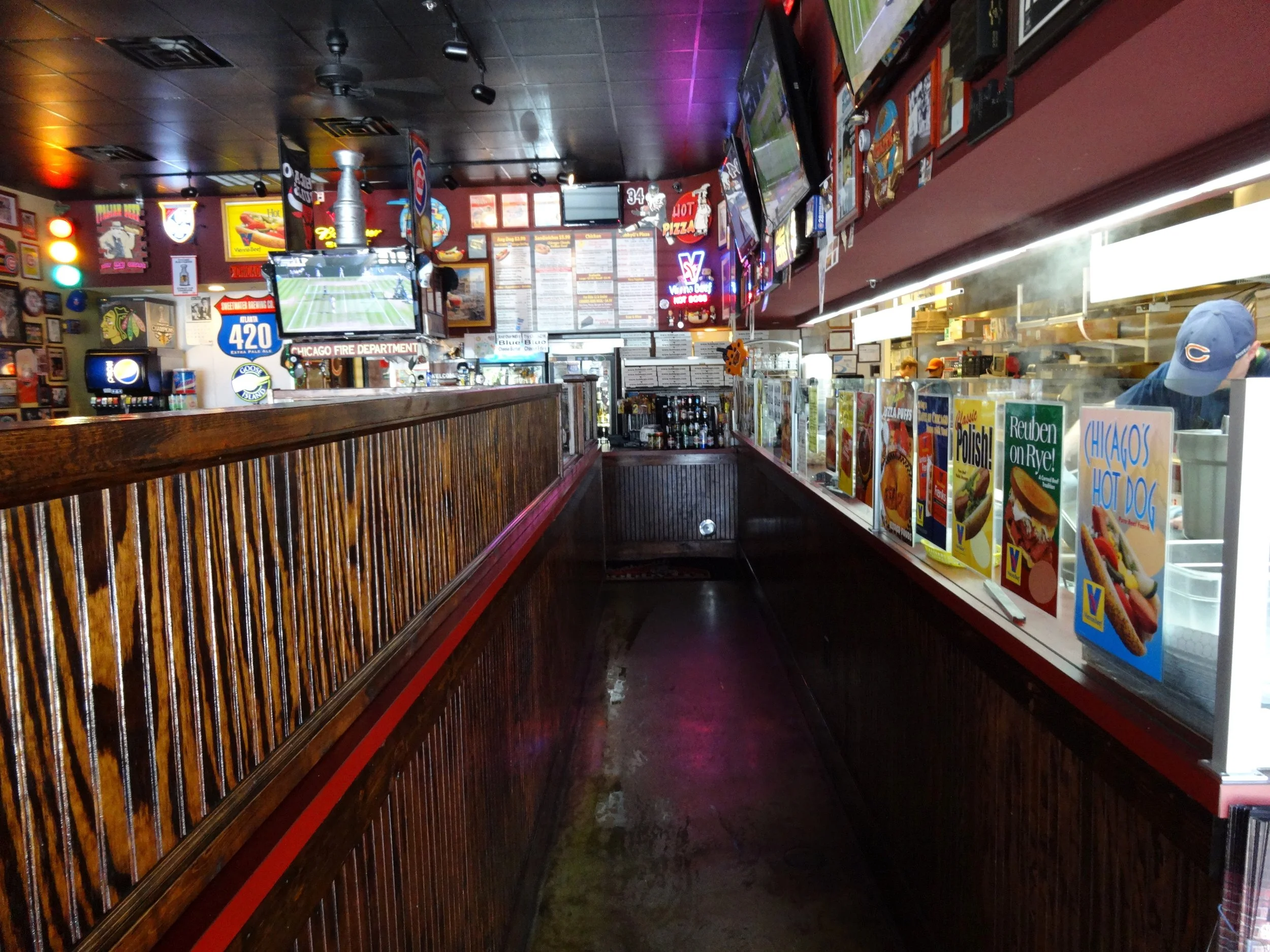 Interior of a sports bar with a wooden counter, multiple TV screens showing sports, colorful neon signs, framed pictures, and a menu display on the wall, along with a counter window with various food advertisements.
