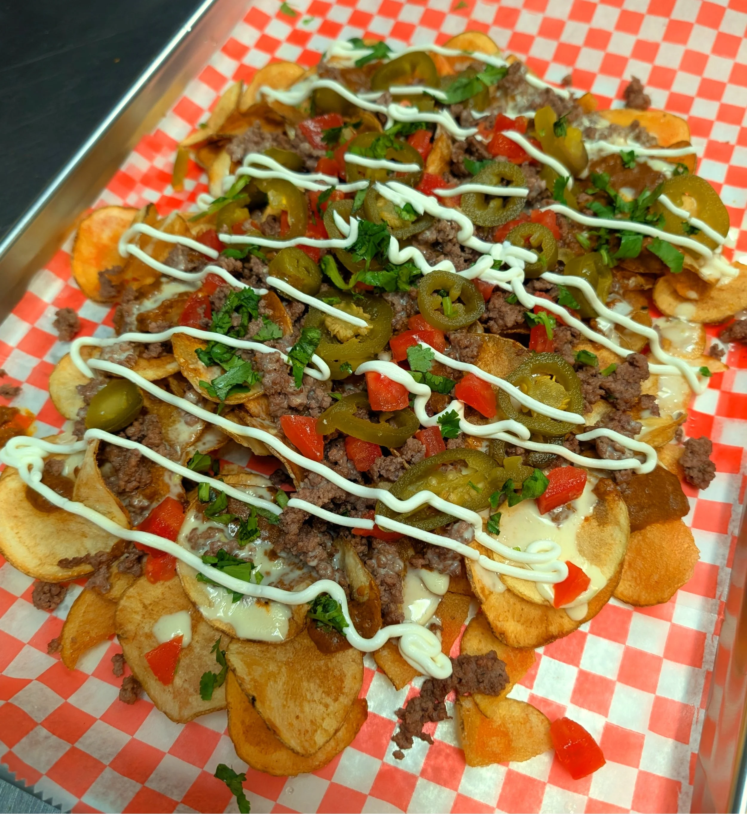 Loaded plate of nachos with potato chips, ground beef, melted cheese, diced tomatoes, jalapeño slices, chopped cilantro, and drizzled white sauce on red checkered paper.