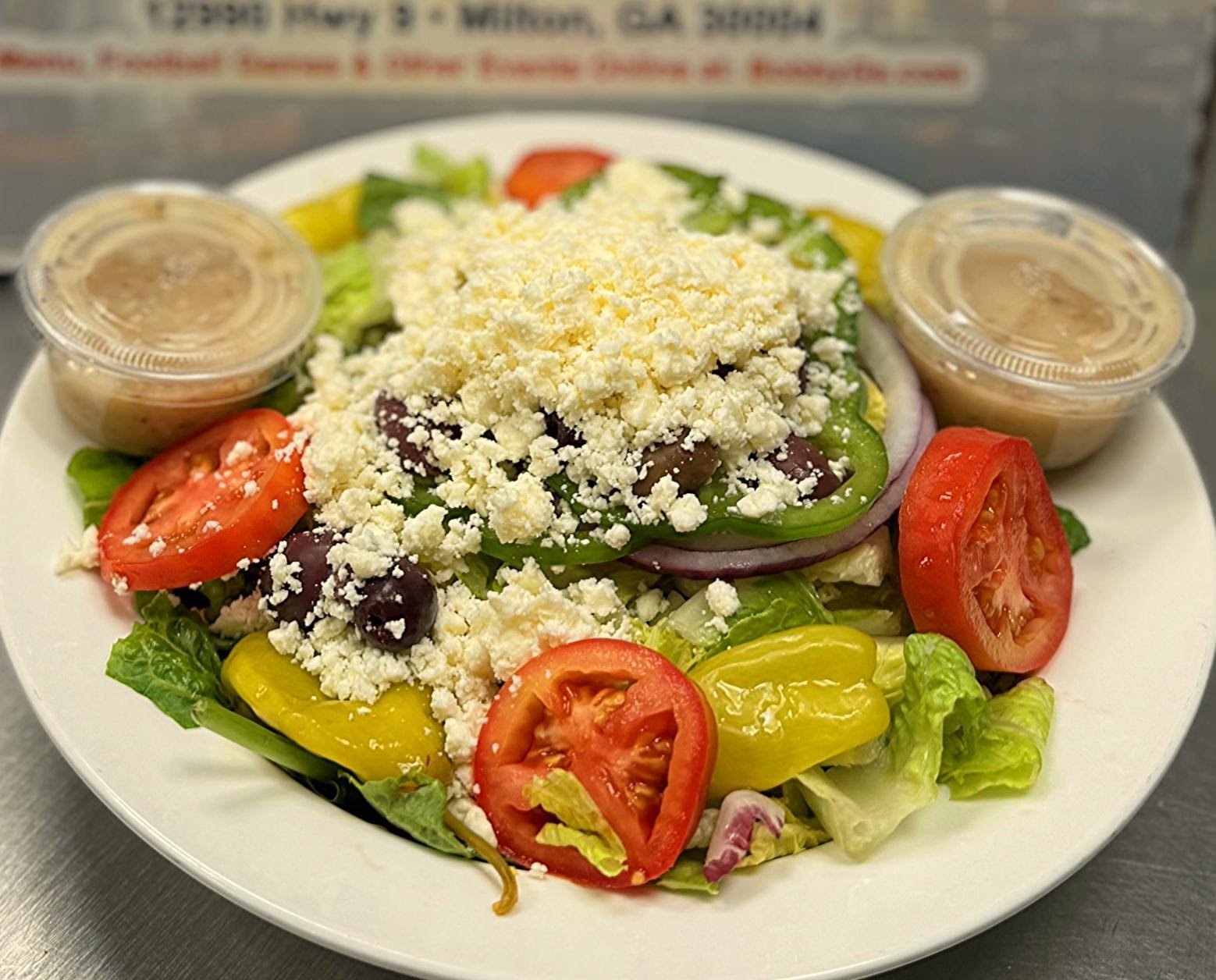 A salad with tomatoes, green bell peppers, lettuce, red onions, black olives, crumbled cheese, and two small containers of dressing.