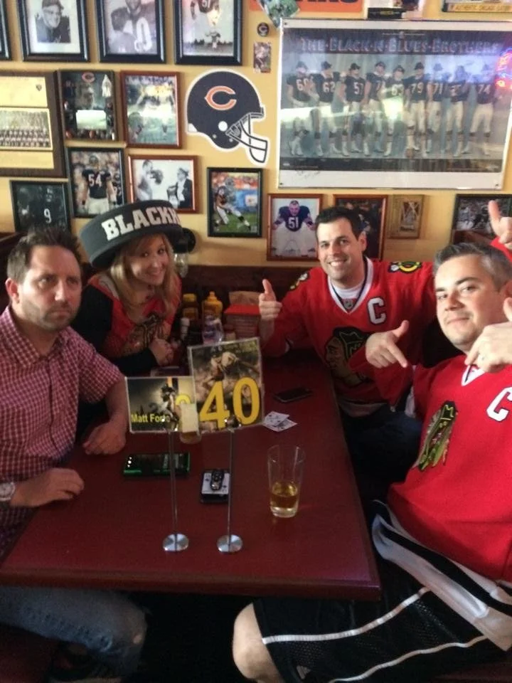 Four people sitting at a table in a sports bar, celebrating with drinks and wearing Chicago Blackhawks hockey jerseys, with sports memorabilia and framed photos on the wall behind them.