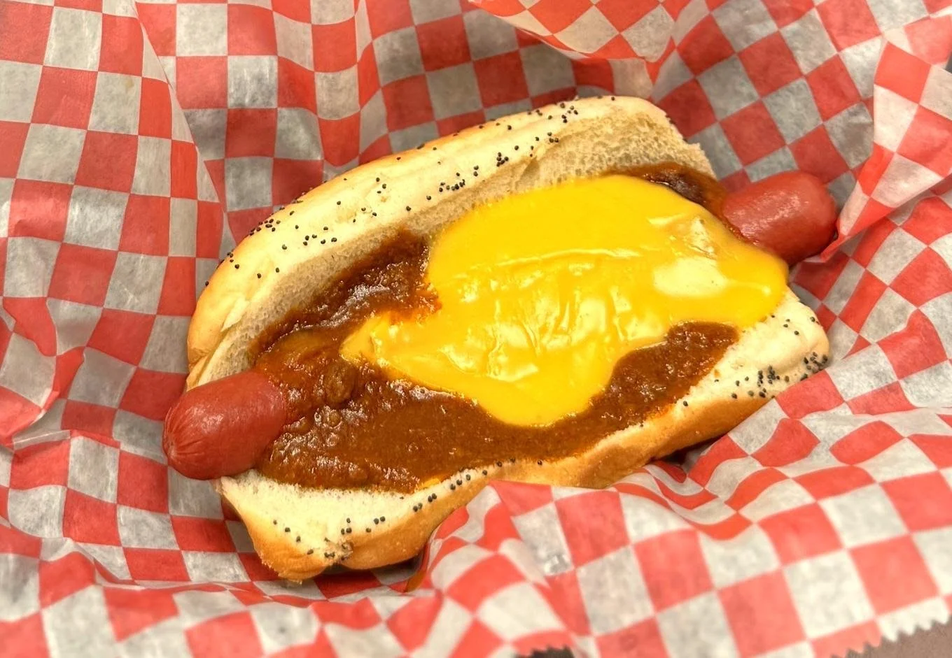 Hot dog in a bun with chili, a folded egg, and two sausages, wrapped in red and white checkered paper.
