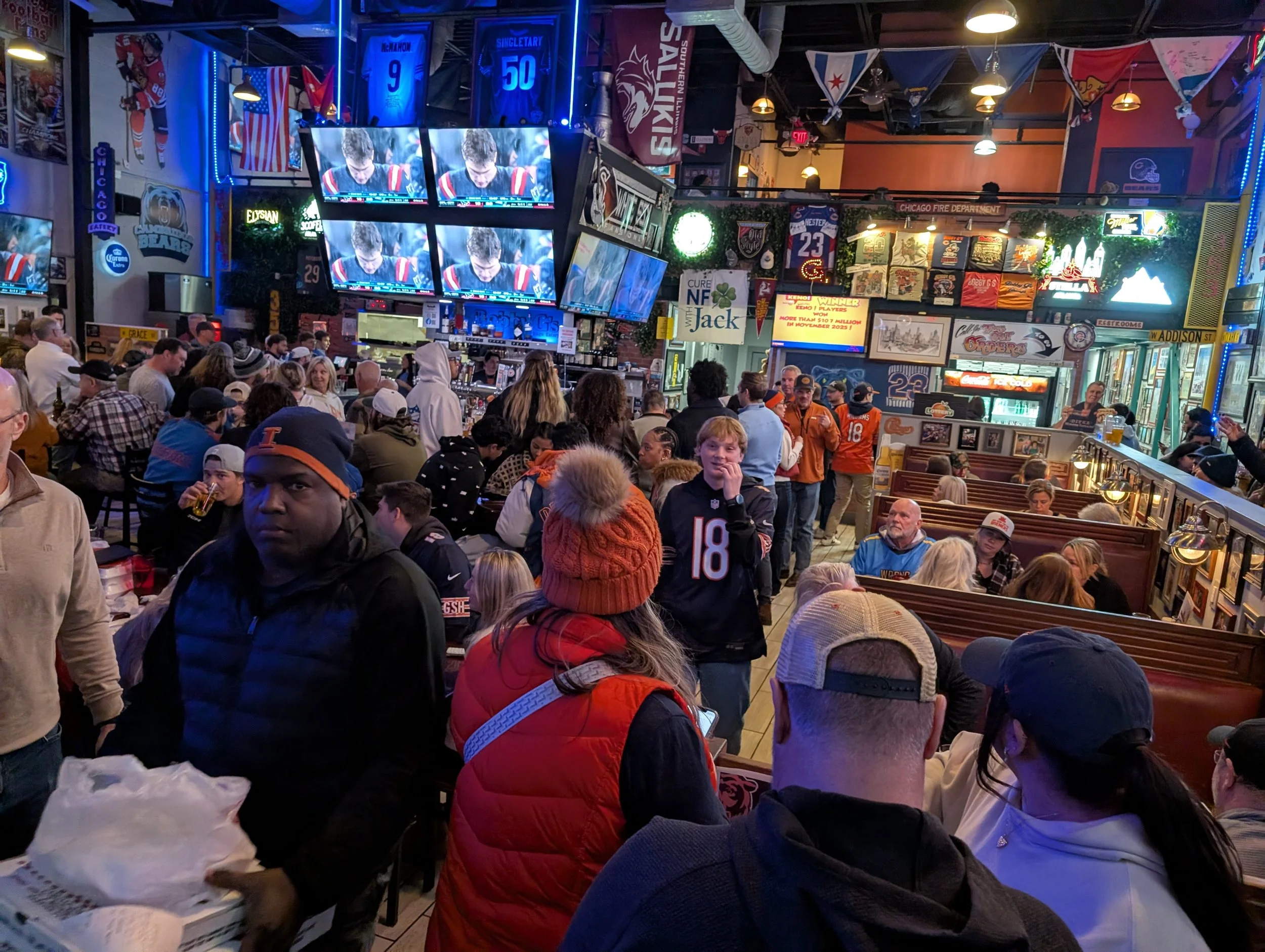 Crowded sports bar filled with people watching multiple screens showing a football game, decorated with sports jerseys, banners, and memorabilia.