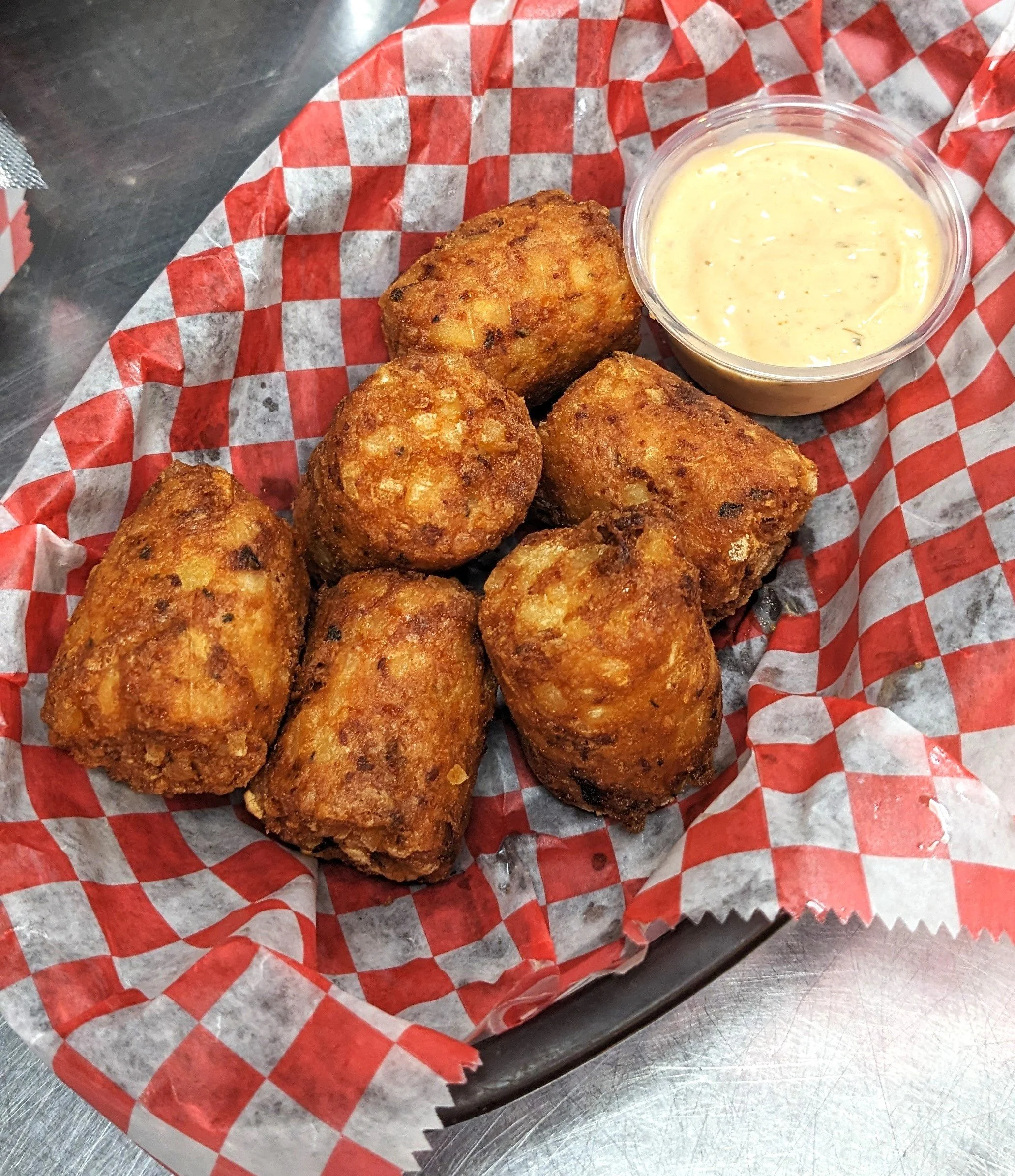 Five fried hush puppies with a side of creamy dipping sauce in a small plastic container on red and white checkered paper in a basket.