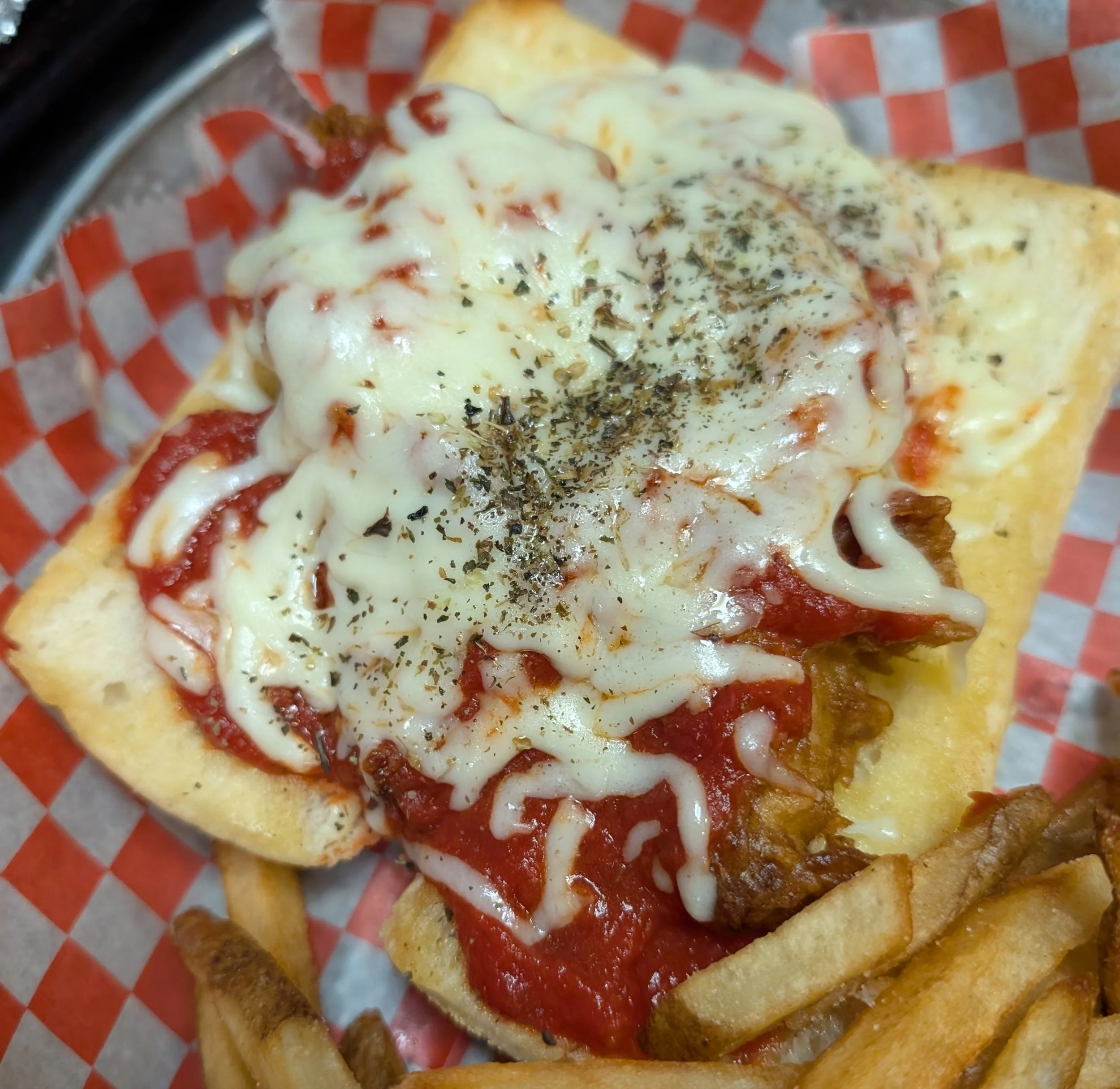 Battered fish fillet topped with melted cheese, tomato sauce, and herbs, served with French fries in a basket lined with red checkered paper.