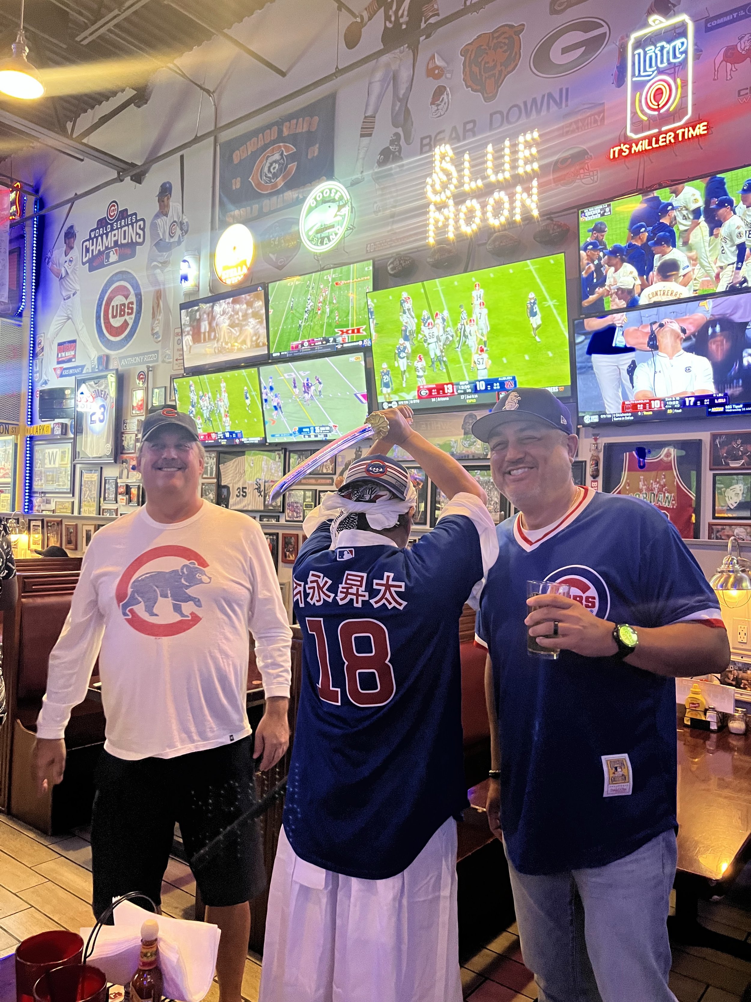 Three men at a sports bar celebrating, with a person in the middle raising a large knife over their head. The bar is decorated with Chicago Cubs memorabilia and multiple television screens showing a baseball game.