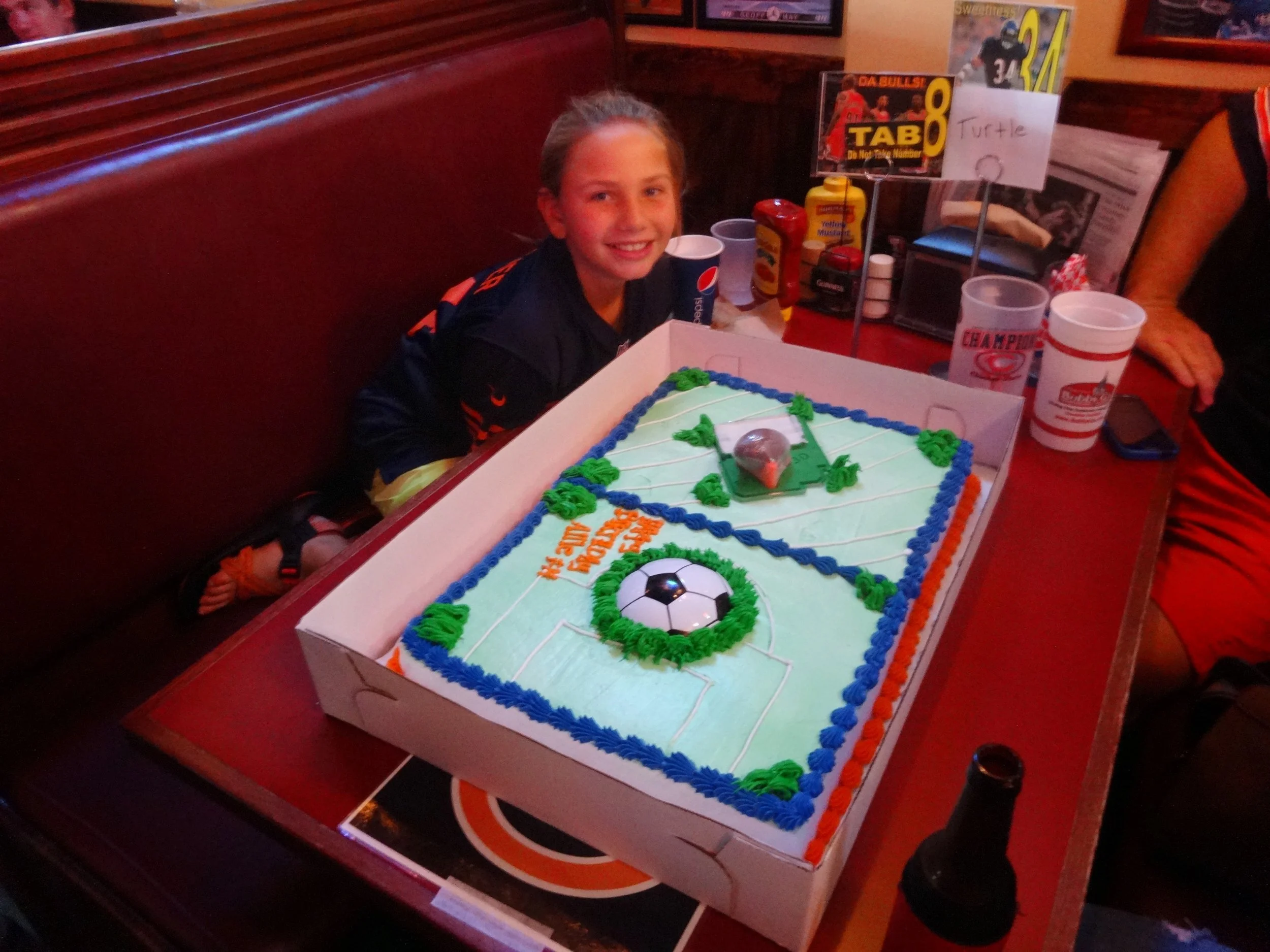 A girl with a big smile sitting at a table with a hockey-themed cake, decorated with green and blue icing, a soccer ball, and a mini hockey puck, celebrating a birthday.