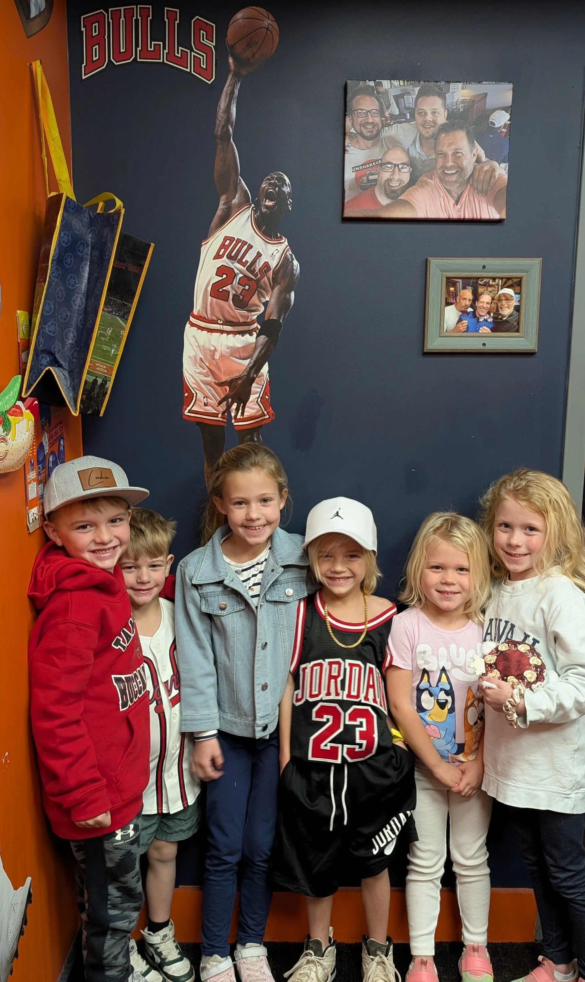 Group of six children standing in front of a wall decorated with an image of Michael Jordan and framed photos, some wearing Chicago Bulls jerseys and hats.