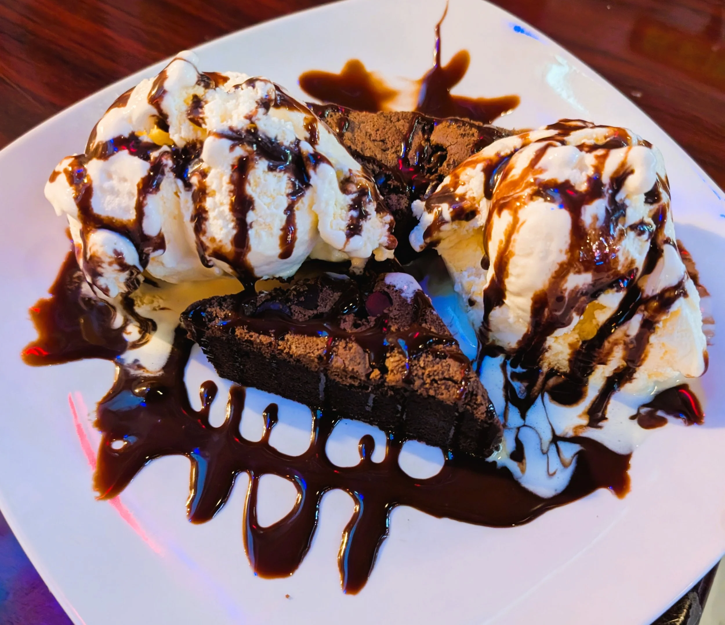 A dessert plate with two scoops of vanilla ice cream topped with chocolate syrup, a slice of chocolate cake garnished with chocolate syrup, and a piece of chocolate brownie, all drizzled with chocolate syrup on a white plate.