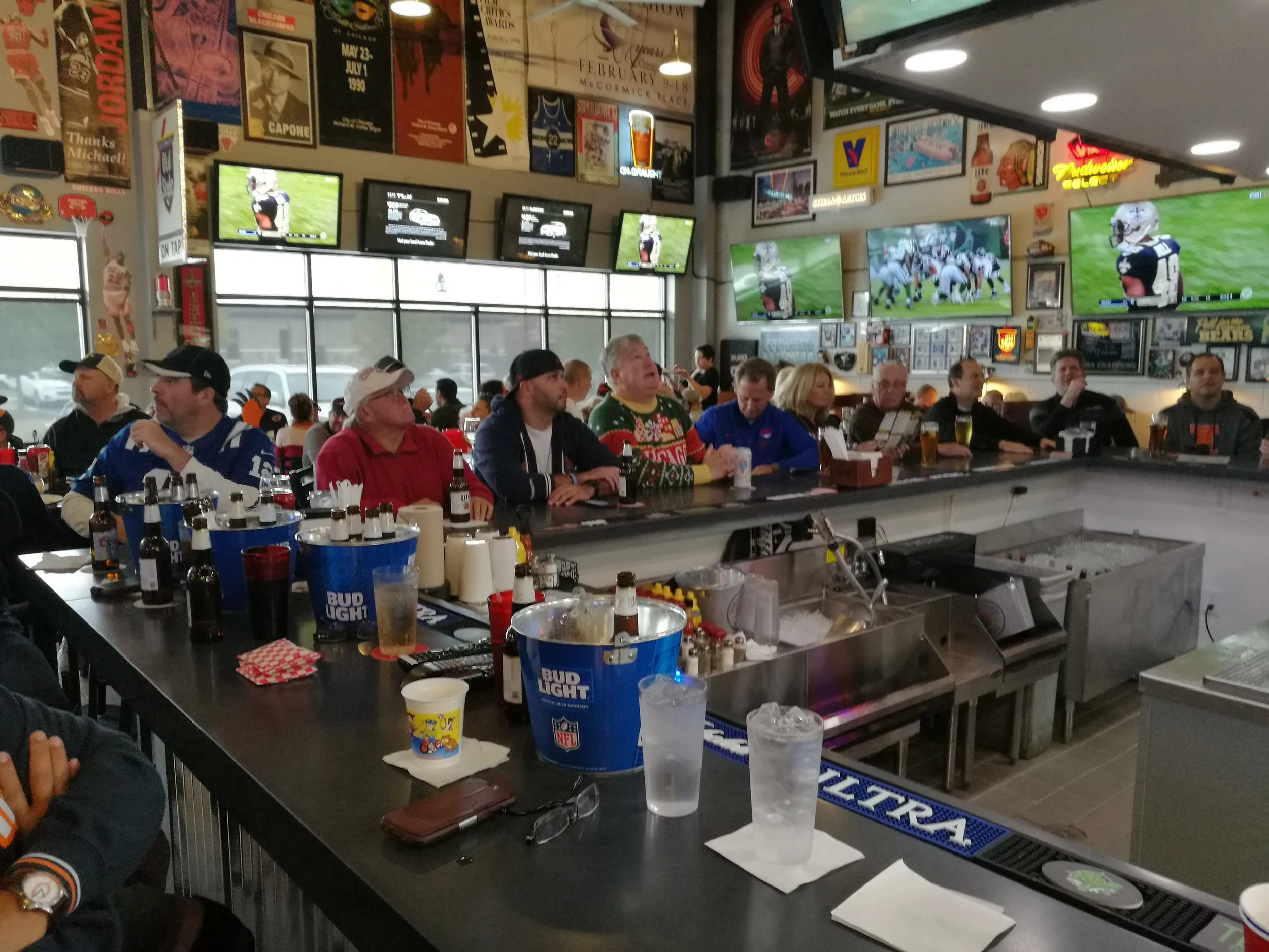 People sitting at a bar in a sports bar, watching multiple TV screens showing football games. The bar table has several drinks, condiment bottles, and buckets of beer. The background displays sports memorabilia and posters.