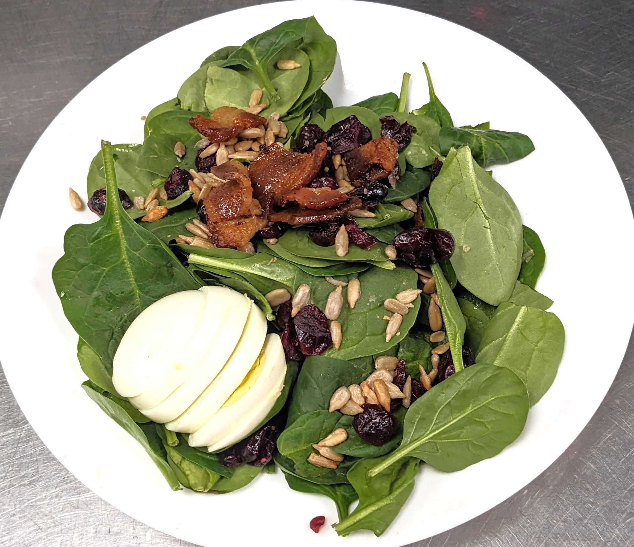 Spinach salad with slices of hard-boiled egg, dried cranberries, sunflower seeds, candied bacon pieces, and crumbled bacon on top.