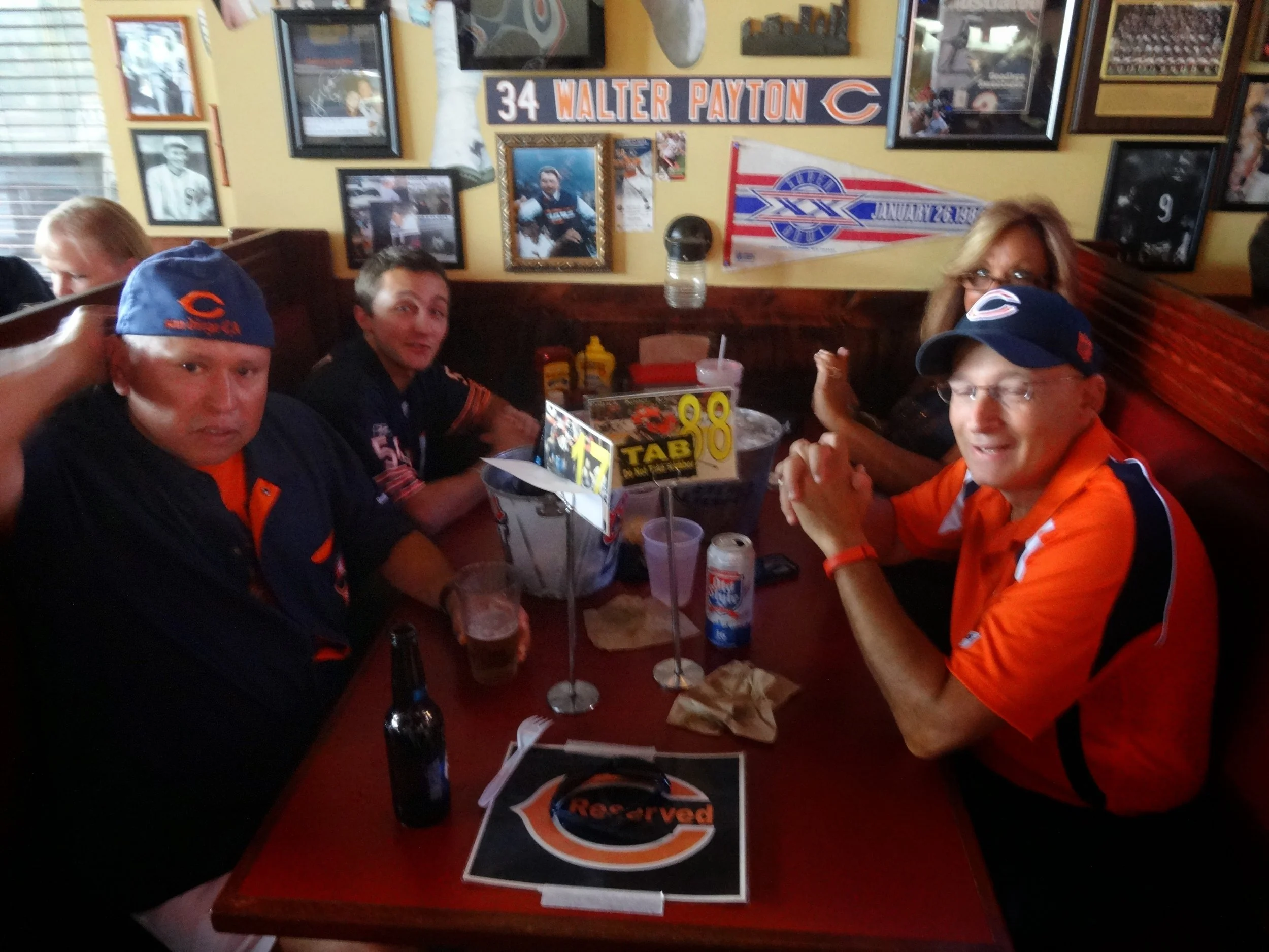 Four people sitting at a table in a sports-themed restaurant decorated with Chicago Bears memorabilia. Two men wear Chicago Bears jerseys and hats, and there is a woman with glasses and long hair. The table has drinks, condiments, napkins, and a rese