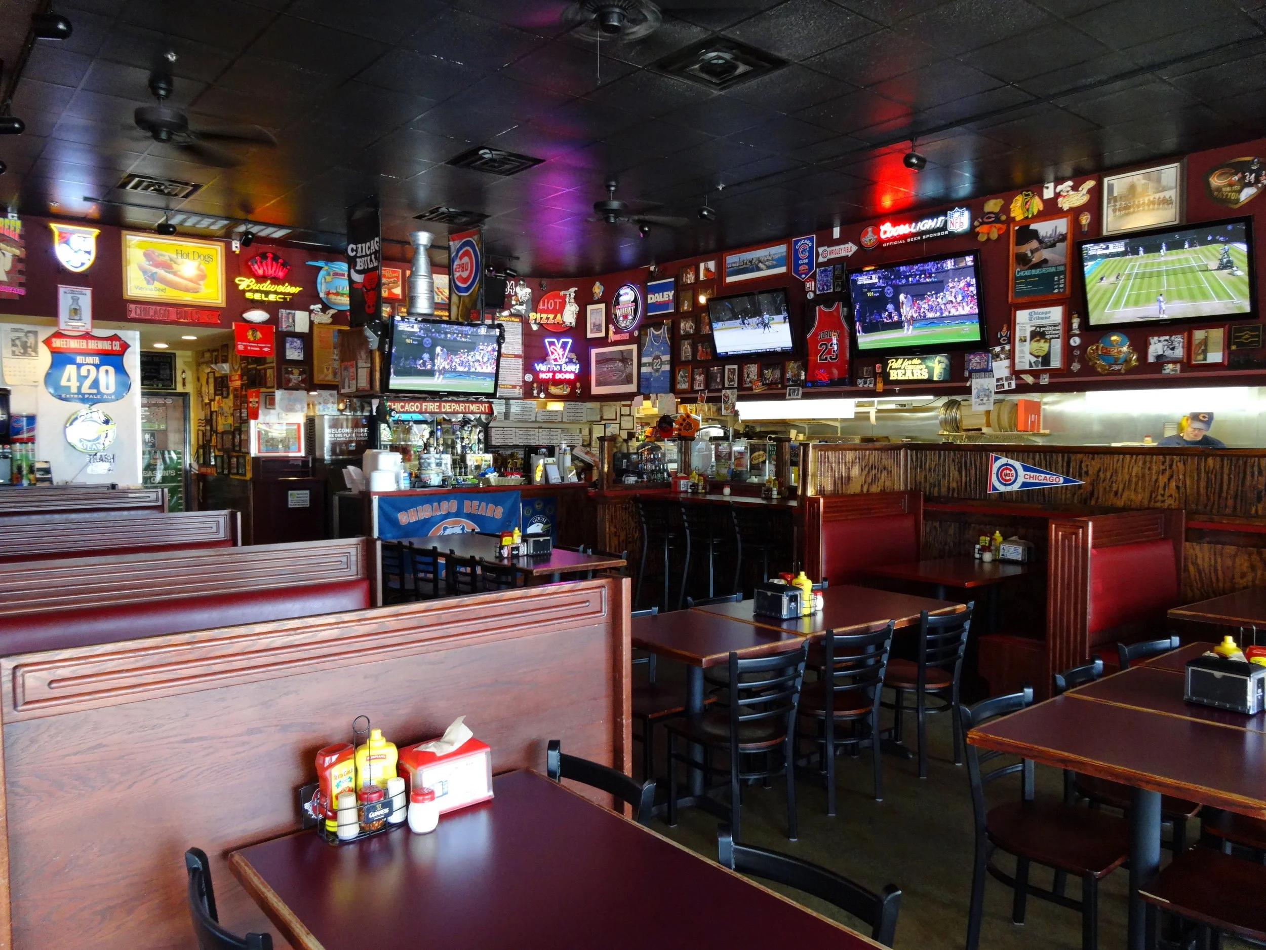Interior of a sports bar with multiple television screens showing a tennis match and other games, wooden tables and chairs, neon signs, sports memorabilia, and condiments on tables