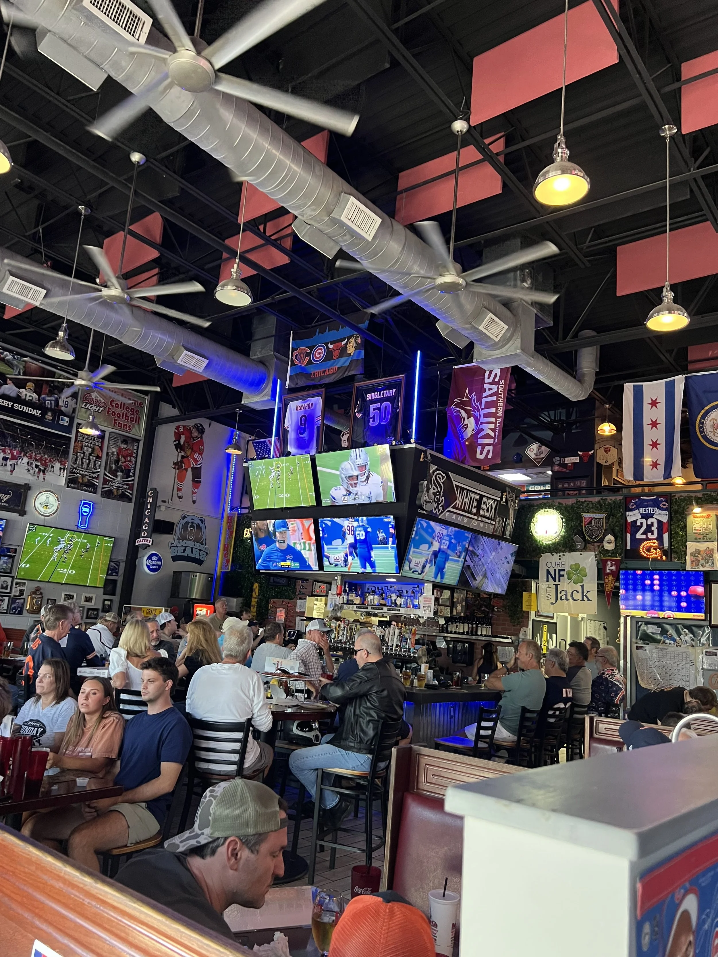 Interior of a sports bar filled with patrons watching multiple large TVs showing football games and sports highlights. Decor includes sports banners, flags, and memorabilia.