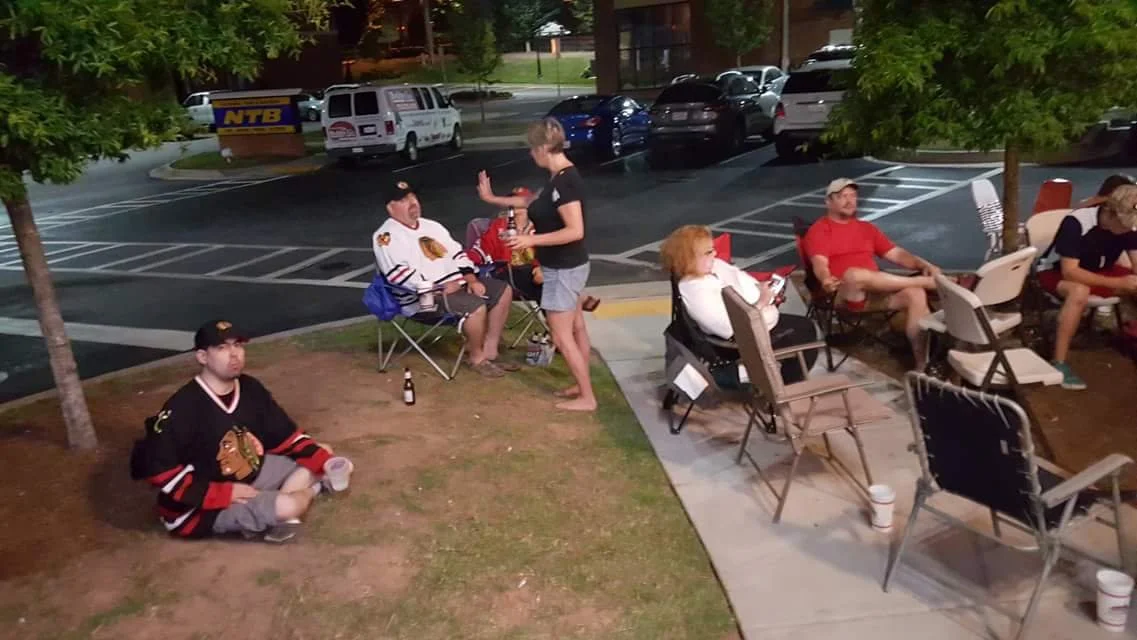 People sitting on chairs and on the ground outside, some wearing hockey jerseys, engaging in conversation and relaxing near a parking lot with cars and trees.