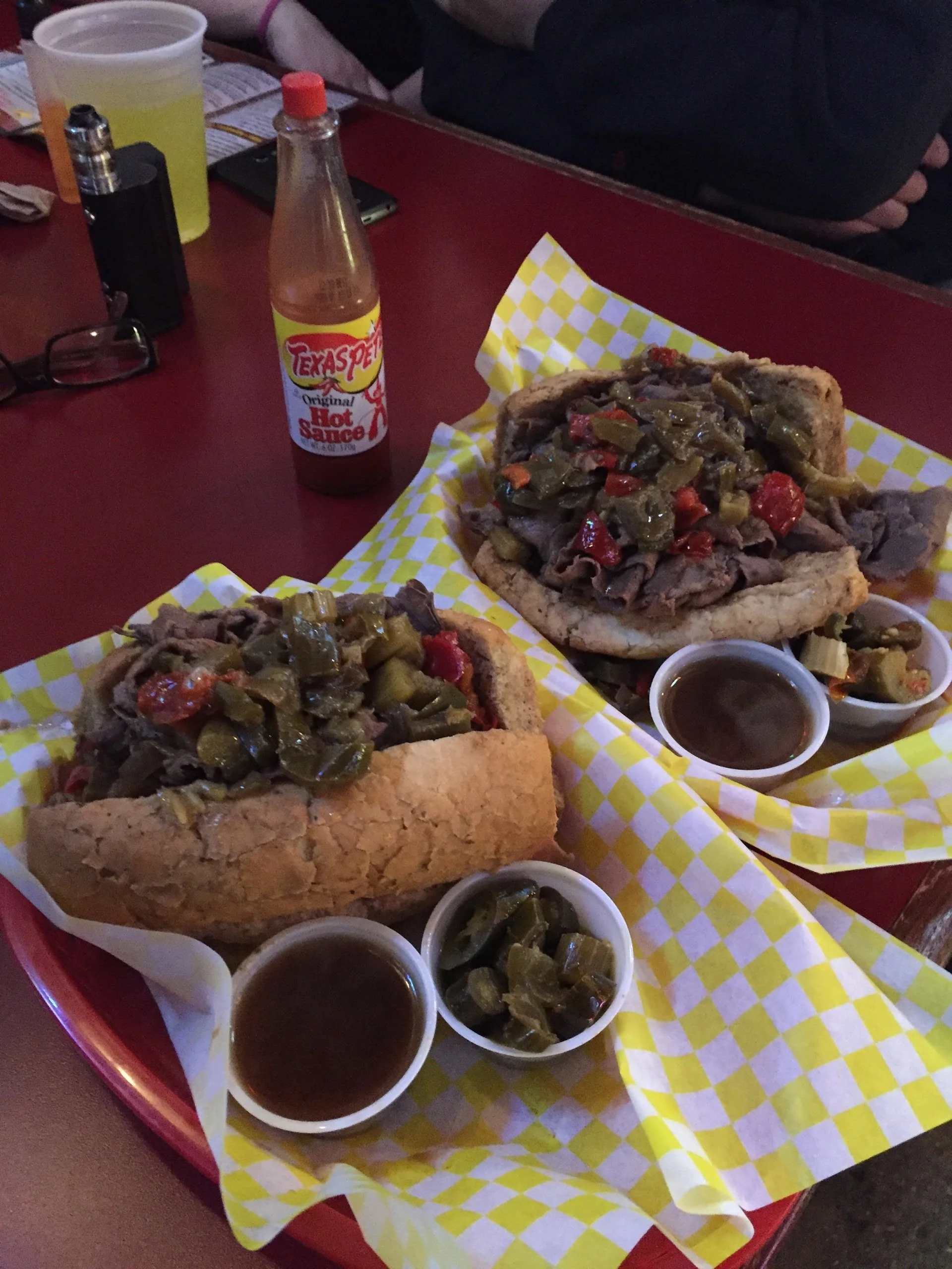 Two hoagies topped with green and red peppers on yellow checkered paper, with small bowls of dark sauce and pickled peppers, and a bottle of Texas Pete hot sauce on a red table.