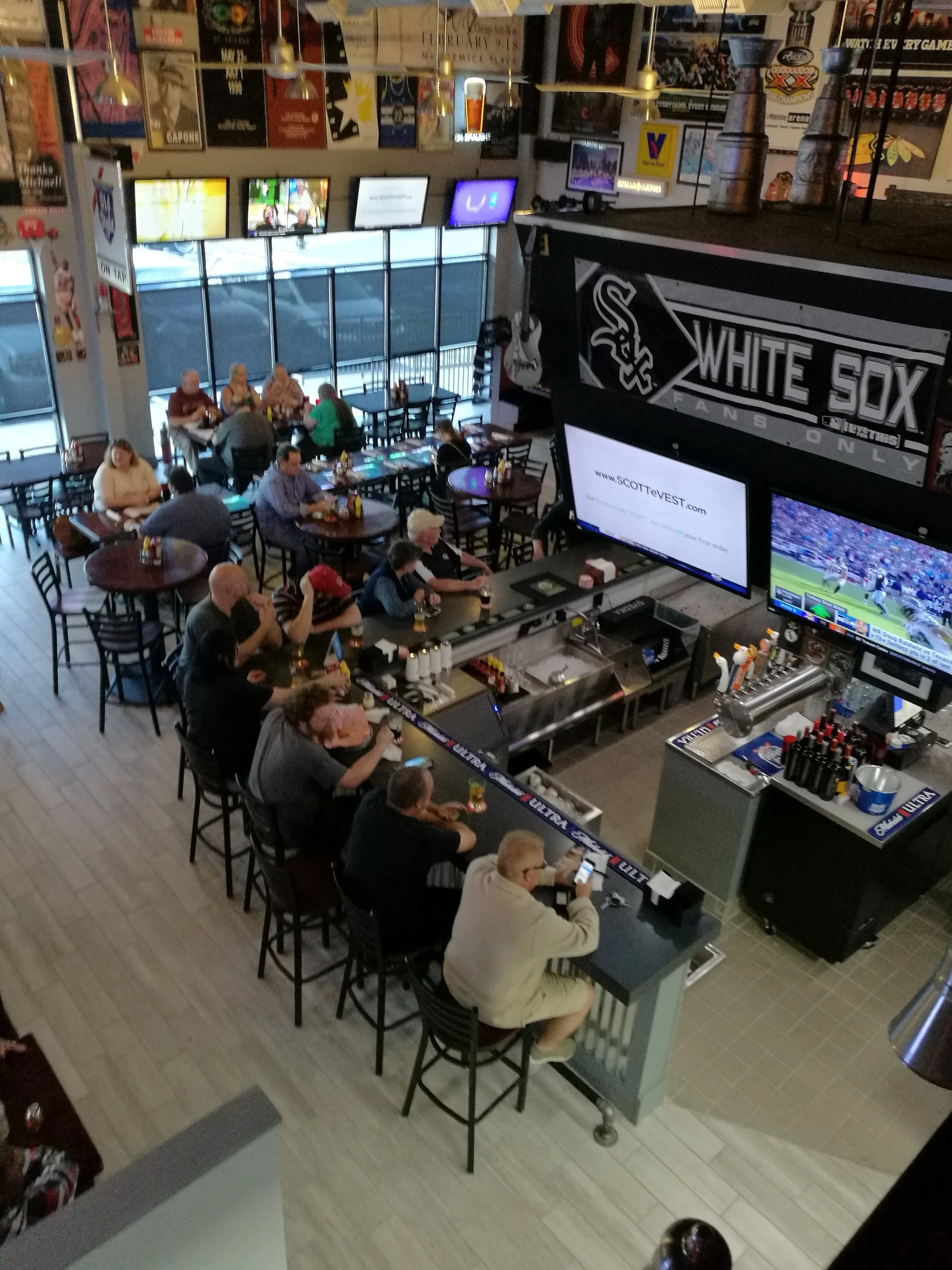 A sports bar with multiple tables occupied by patrons, some watching games on large TVs, with a bar area in the foreground. Decor includes sports memorabilia and a large Chicago White Sox banner.