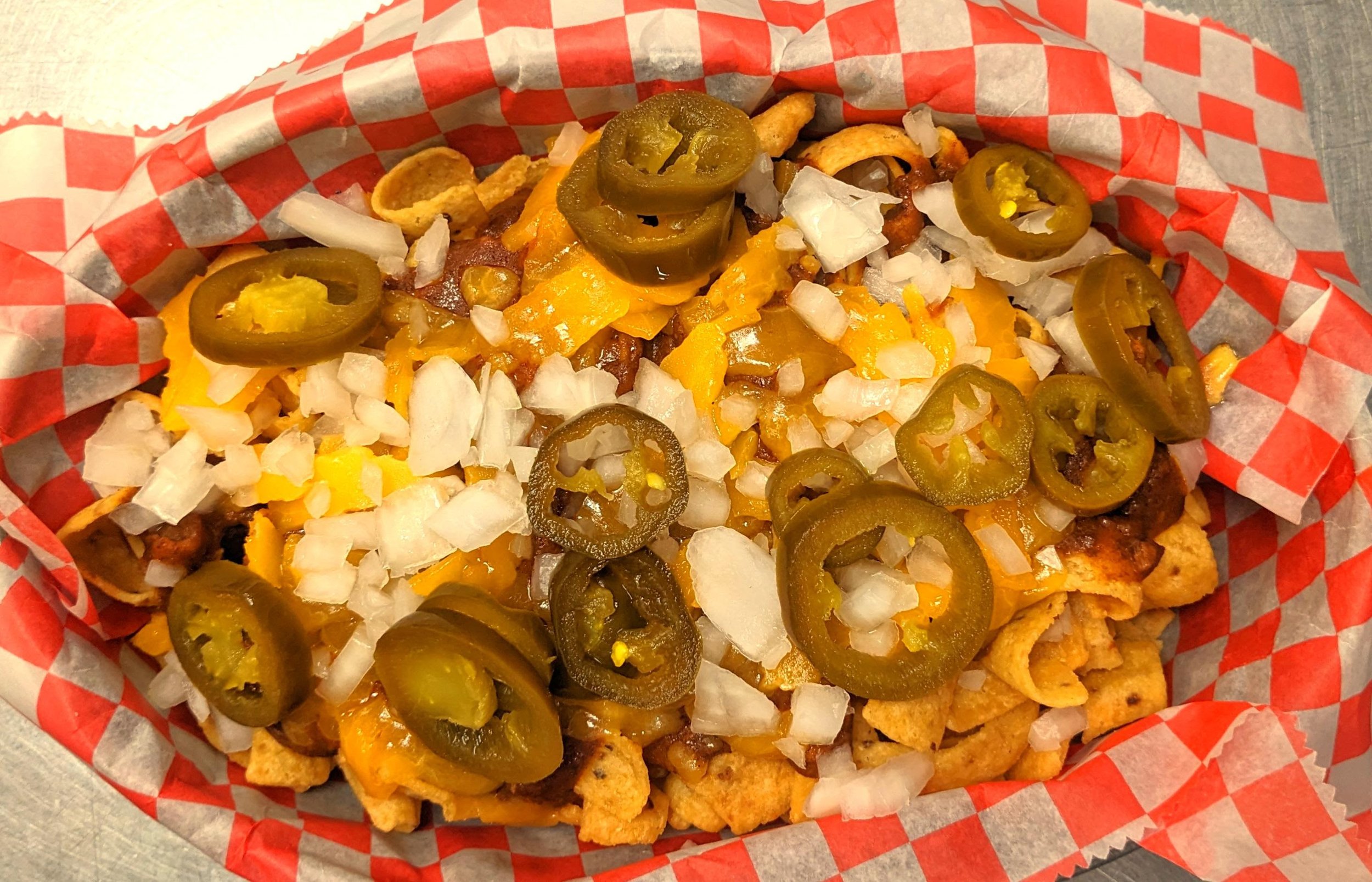 Close-up of a plate of loaded nachos with jalapenos, shredded cheese, diced onions, and tortilla chips in a basket lined with red and white checkered paper.