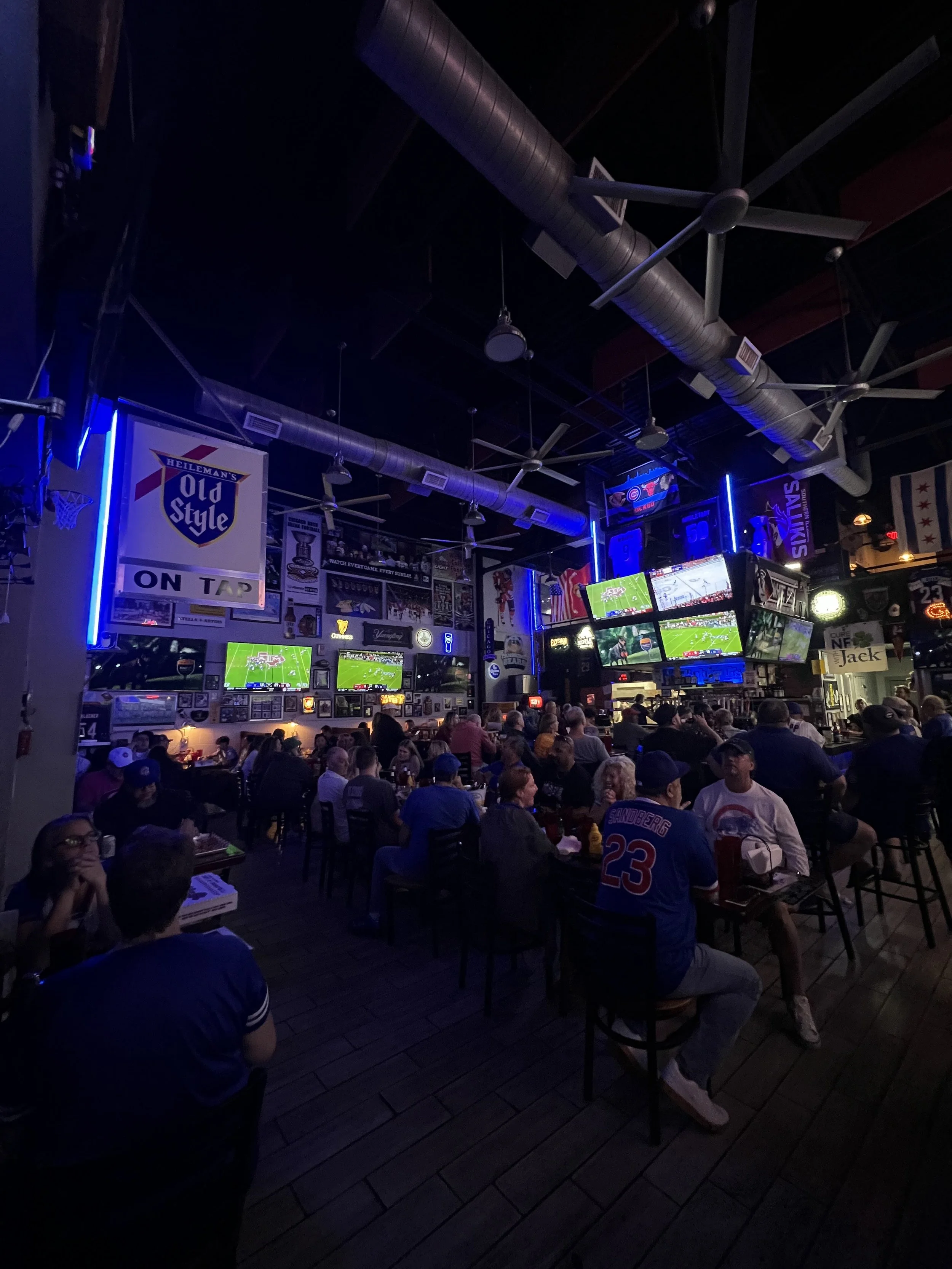 A crowded sports bar with many people watching multiple television screens showing a football game, decorated with sports memorabilia and neon signs.