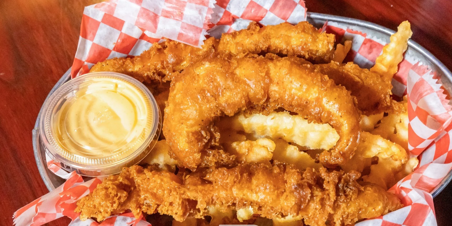 Fried chicken tenders with a side of creamy dipping sauce in a clear cup, served in a metal basket lined with red and white checkered paper.