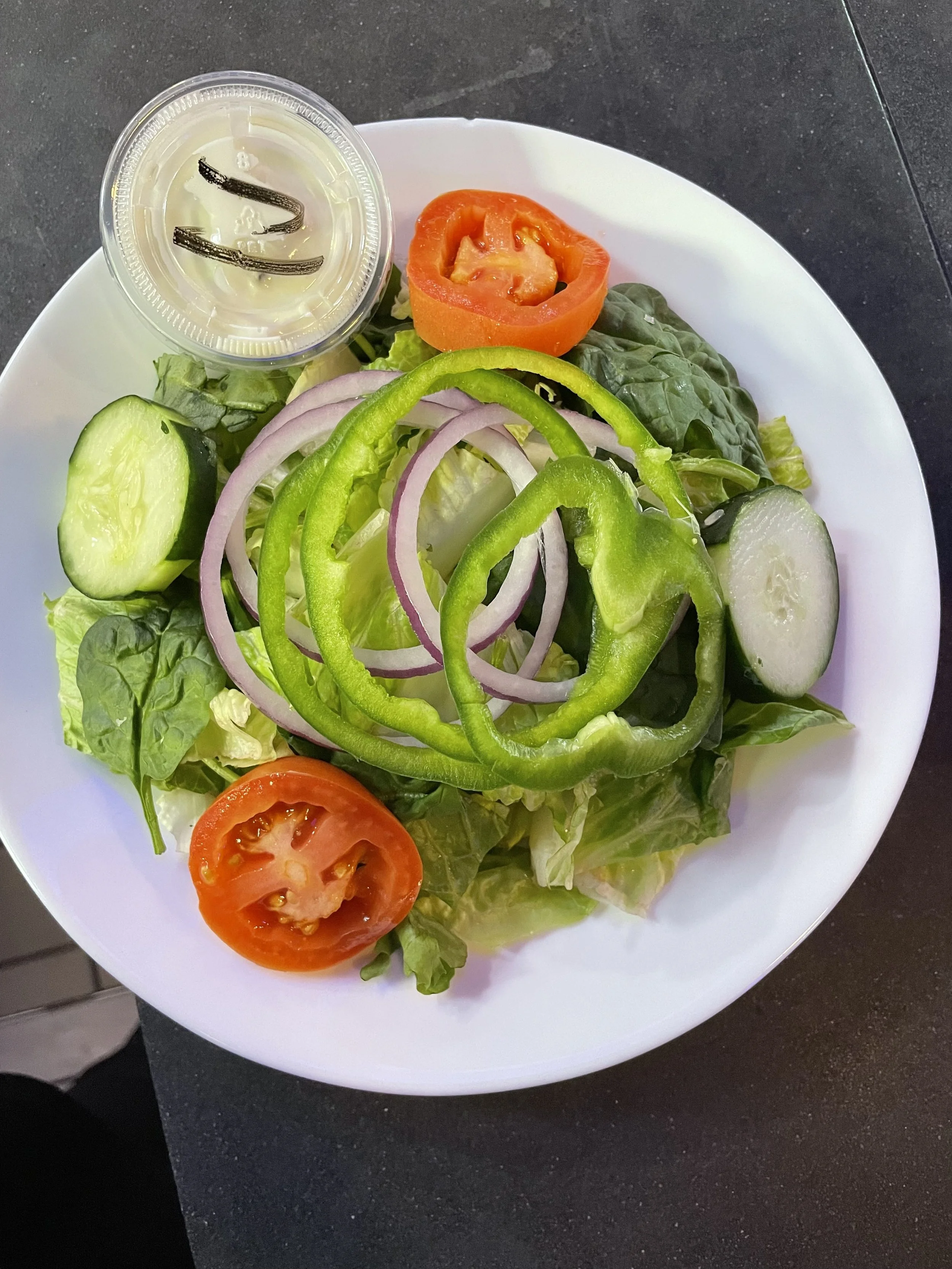 Mixed salad with cucumber, tomato, onion, and green pepper on a white plate, with a small cup of salad dressing on the side.