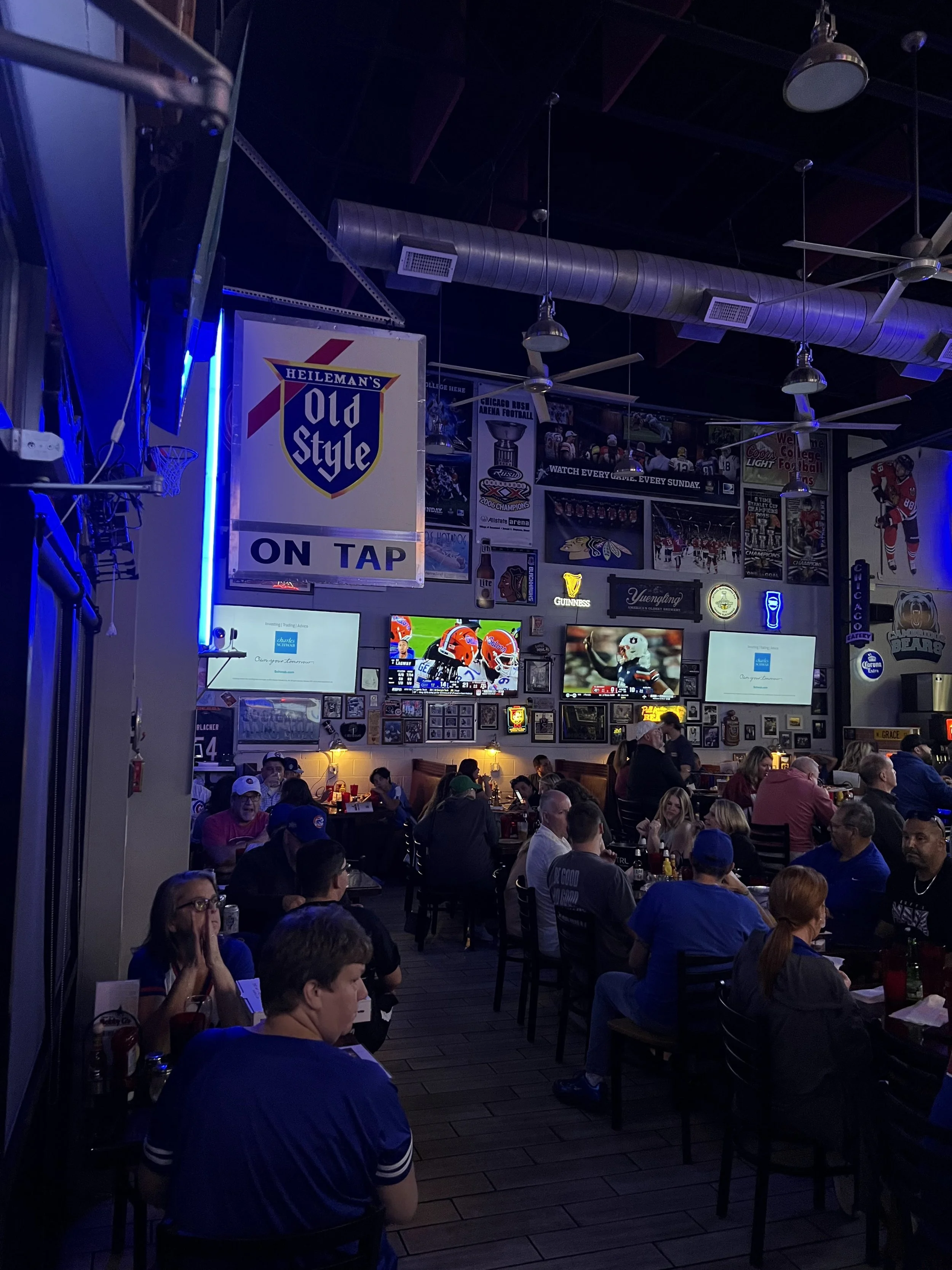 Inside a sports bar with patrons watching football on multiple TVs, decorated with sports memorabilia and neon signs.