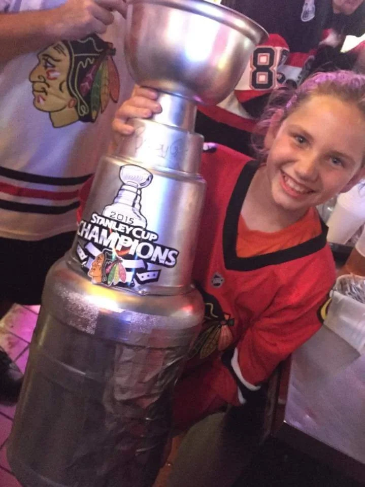 A young girl with blonde hair and a red hockey jersey is smiling and holding the Stanley Cup trophy after winning the 2015 Stanley Cup Finals.