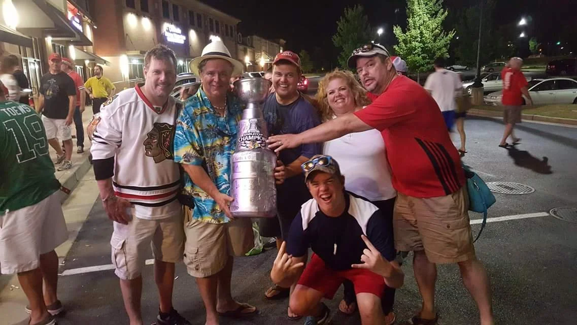 Group of six people standing outside at night, holding the Stanley Cup hockey trophy, celebrating, some making gestures, smiling, in a parking lot with shops in the background.