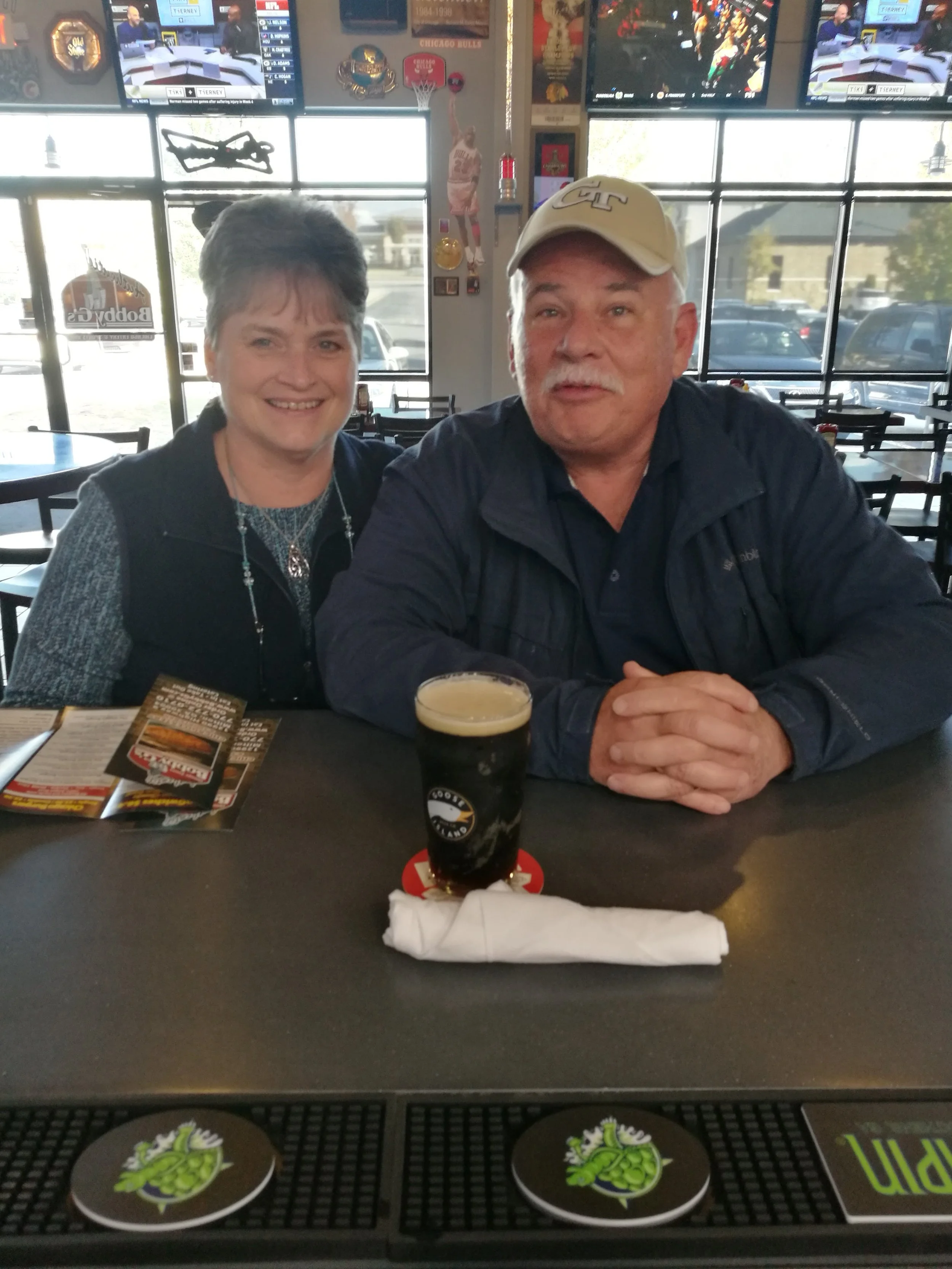 A man and a woman are sitting together inside a restaurant. The man is wearing a beige cap and a dark jacket, while the woman has short hair and is wearing a black vest. There is a pint of dark beer with a foamy head in front of them on the table. Th