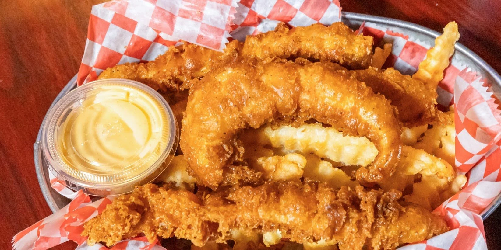 Basket of fried chicken tenders with a side of dipping sauce on a red checkered paper liner.
