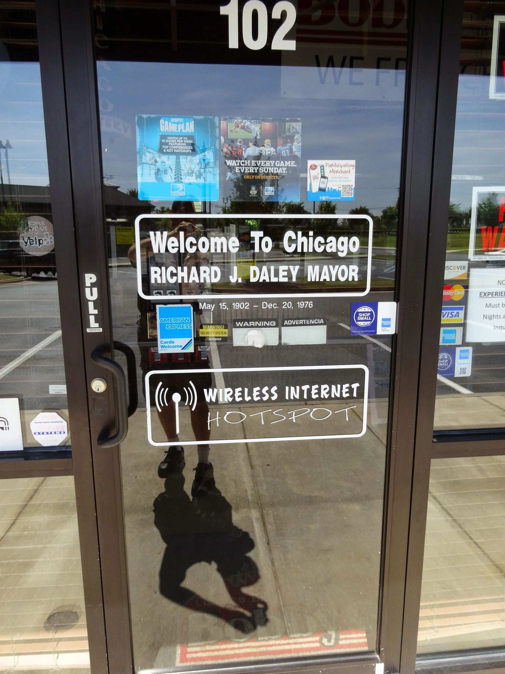 The glass door has signs welcoming visitors to Chicago dedicated to Mayor Richard J. Daley, with his birth and death dates, May 15, 1902 - Dec. 20, 1976. The door also indicates the availability of wireless internet and a hotspot. There are various p